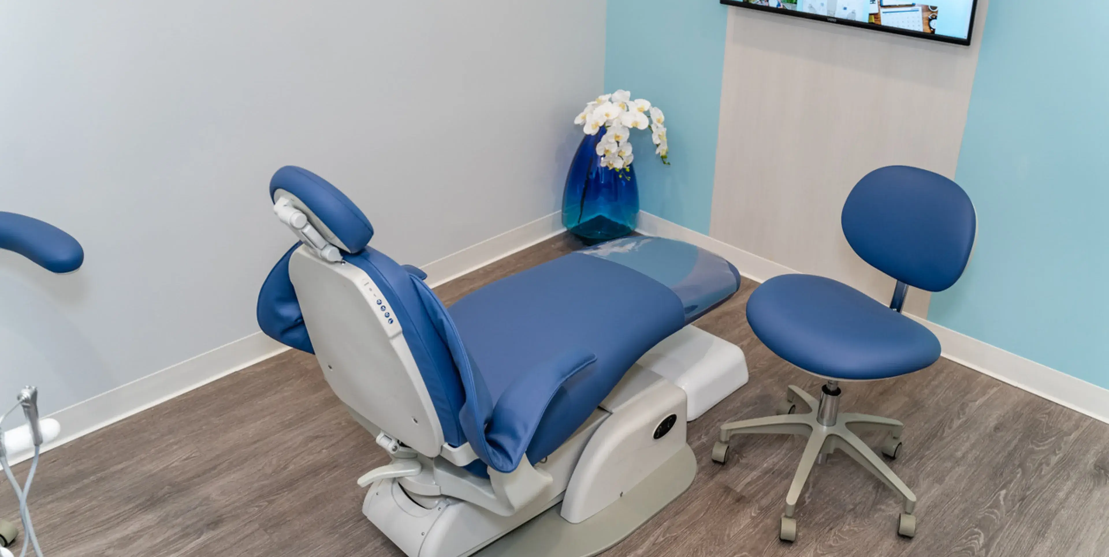 Modern dental clinic room with blue dental chair, matching dentist stool, wood flooring, and a blue vase with white flowers in the corner.