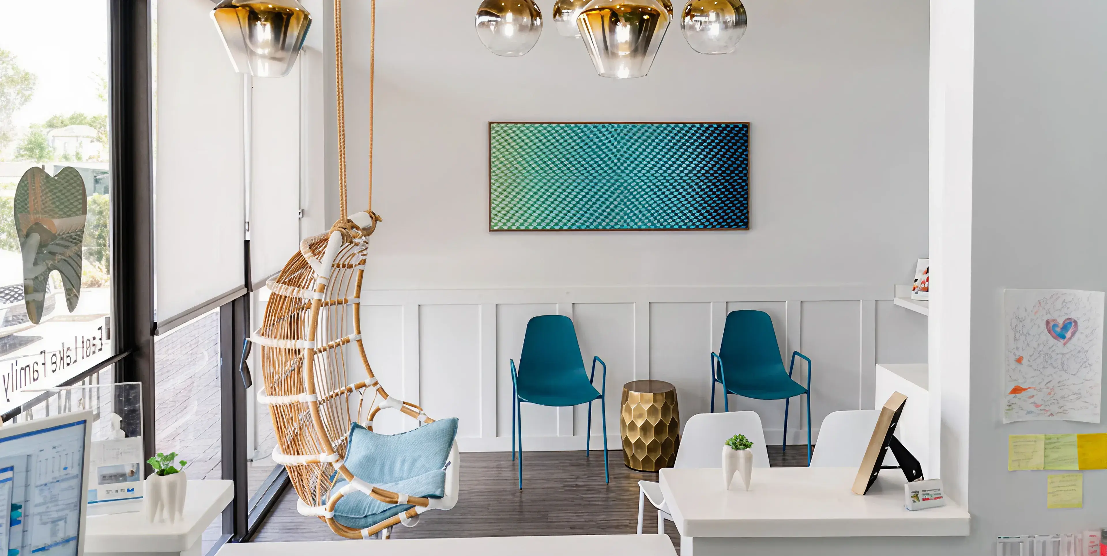 Modern waiting room with wicker hanging chair, two blue chairs, a gold side table, and abstract wall art.