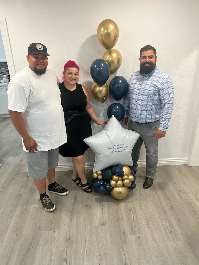 Three people smiling and standing indoors next to a balloon arrangement with gold, blue, and a large silver star-shaped balloon.