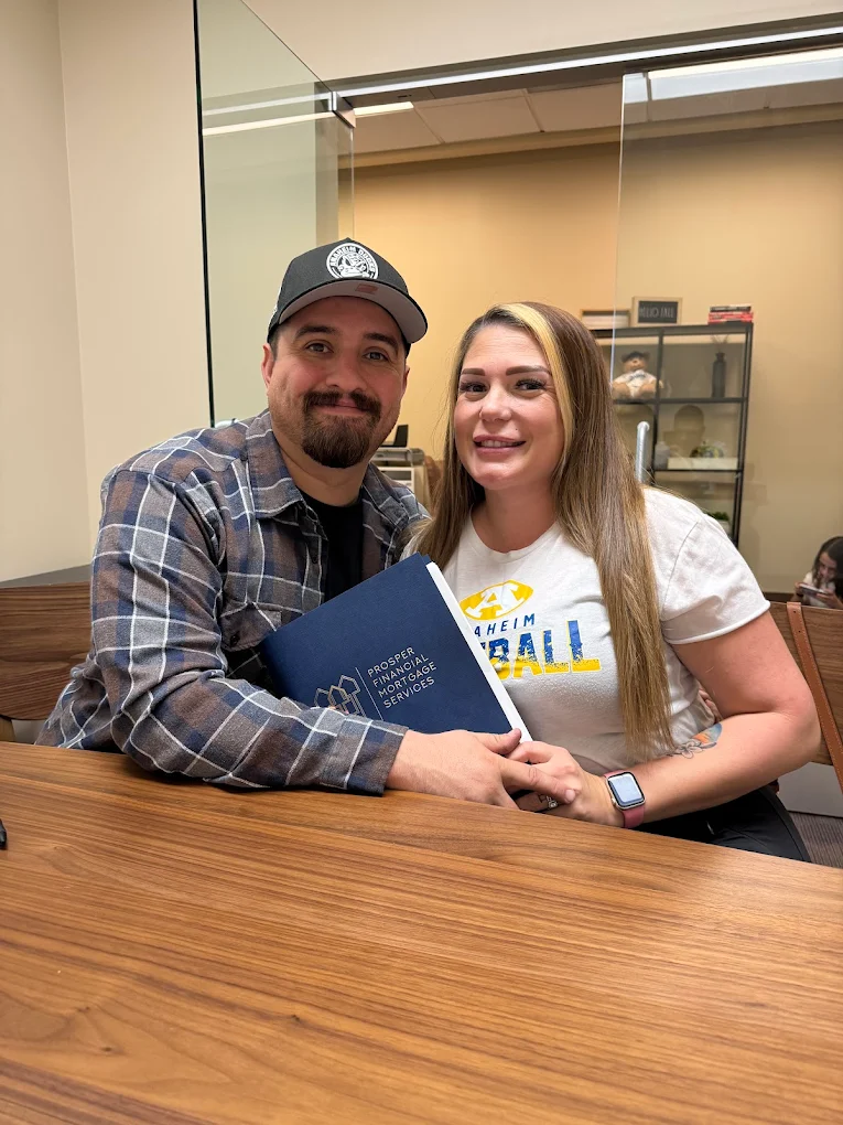 Smiling couple sitting at a wooden table holding a blue folder labeled 'Prosper Financial Mortgage Services' in an office setting.