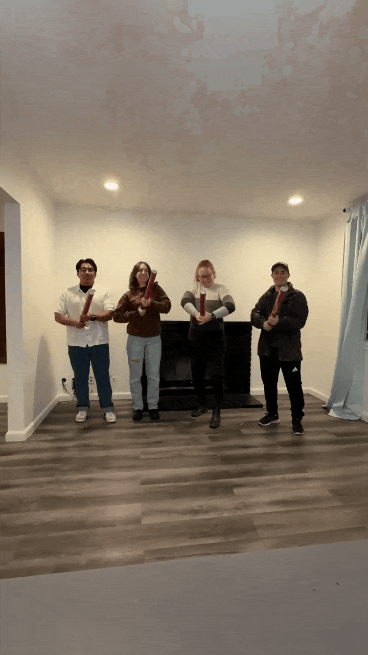 Four people standing indoors holding rolled posters, smiling and moving them playfully in front of a fireplace.