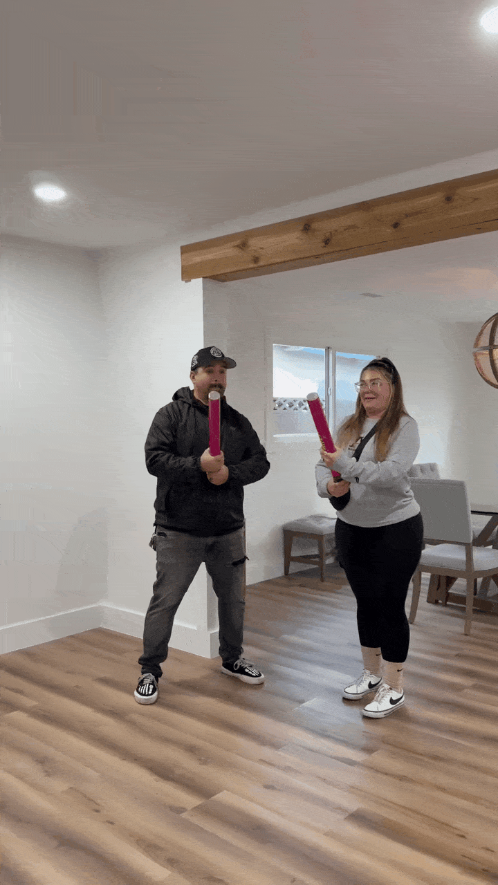 Two people indoors holding and swinging bright pink foam batons playfully on a wood floor with white walls and exposed wooden ceiling beam.