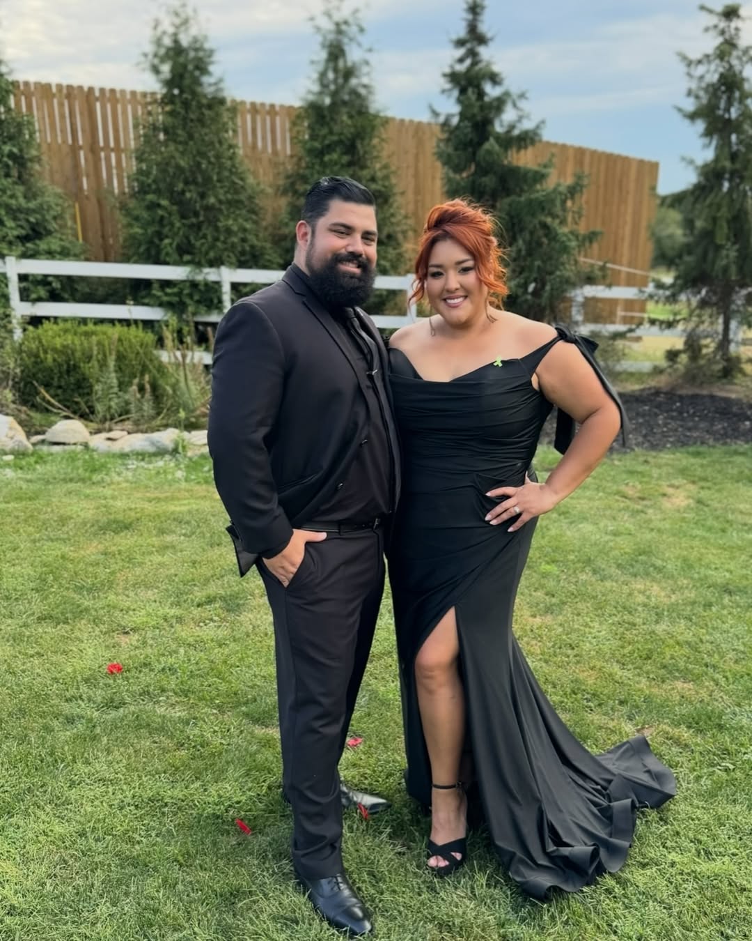 Smiling couple dressed in elegant black attire posing on green lawn with wooden fence and trees in background.