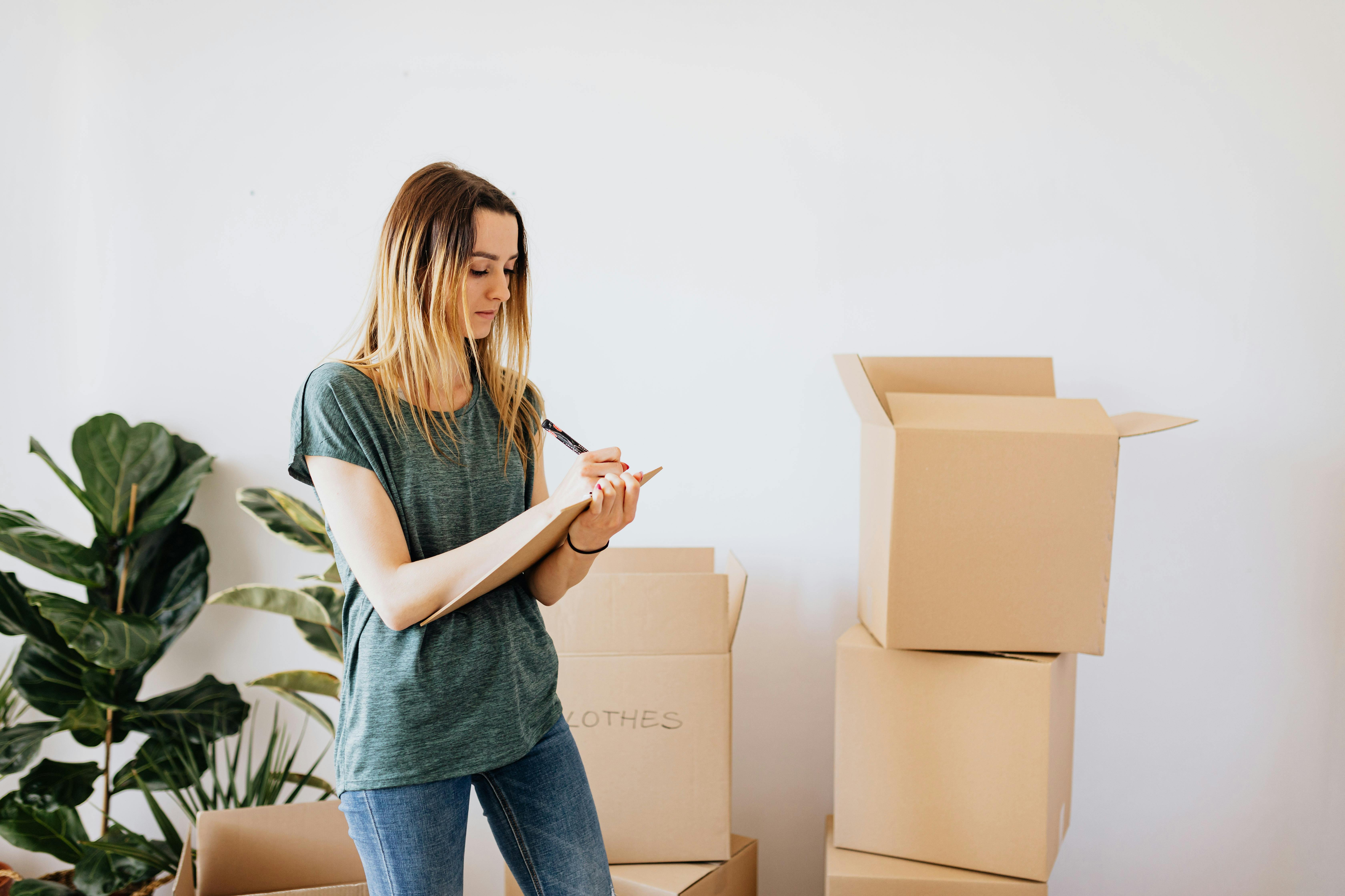 Person standing near stacked cardboard boxes while writing on a clipboard, with houseplant and blank wall behind.