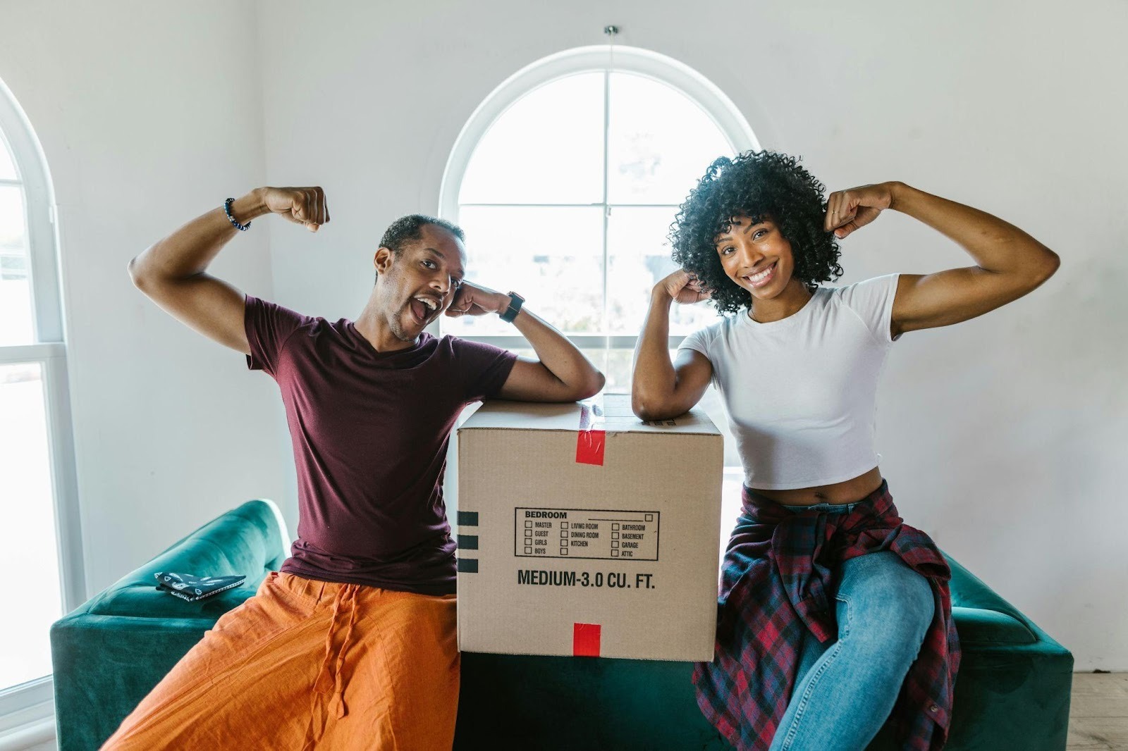 Two people posing beside a moving box on a green couch in a bright, mostly empty apartment with arched windows.