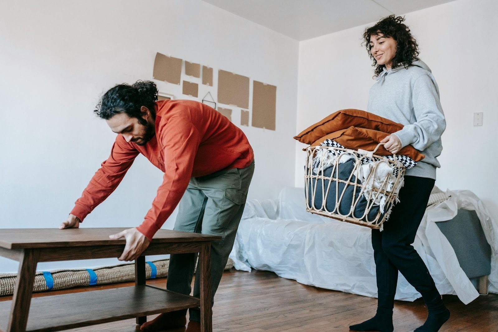 A person sliding a wooden coffee table across hardwood floors while carrying a basket of pillows in a partially furnished living room.
