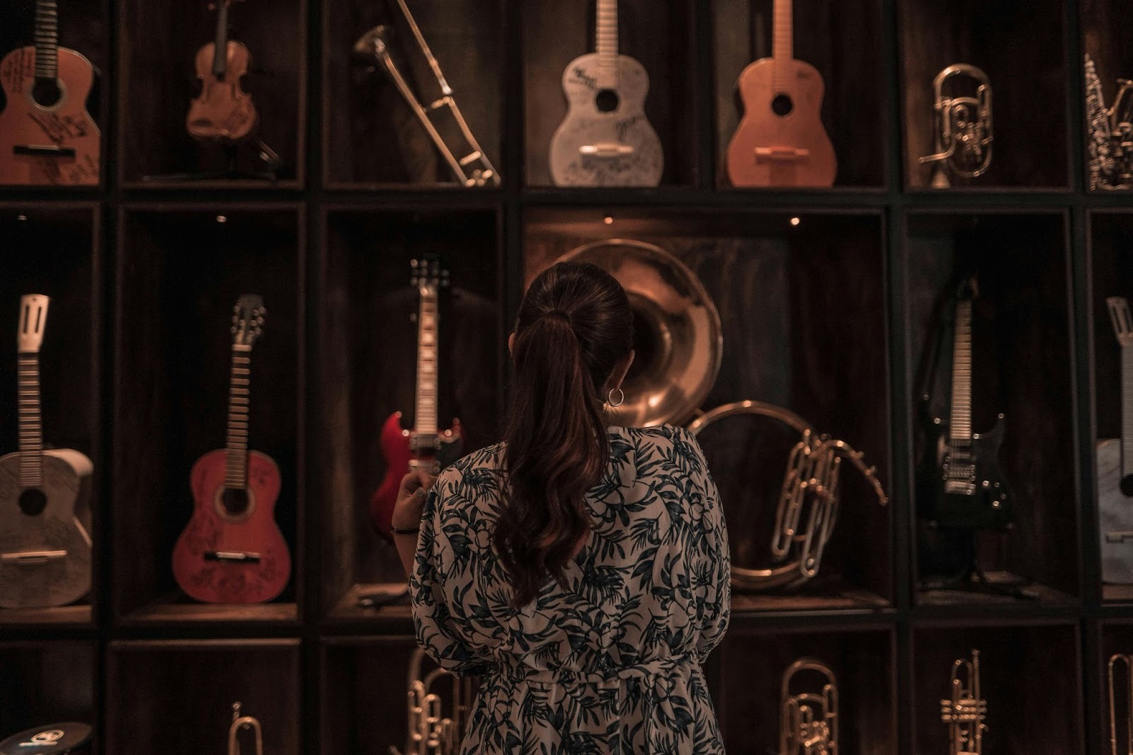 A woman with a ponytail looking at a collection of guitars and brass instruments displayed on a dark wooden shelf.