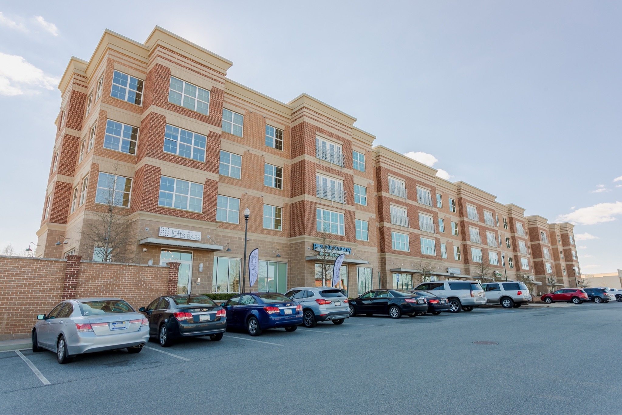 A multi-story brick apartment building standing behind a paved parking lot filled with cars at Lofts at Bass.