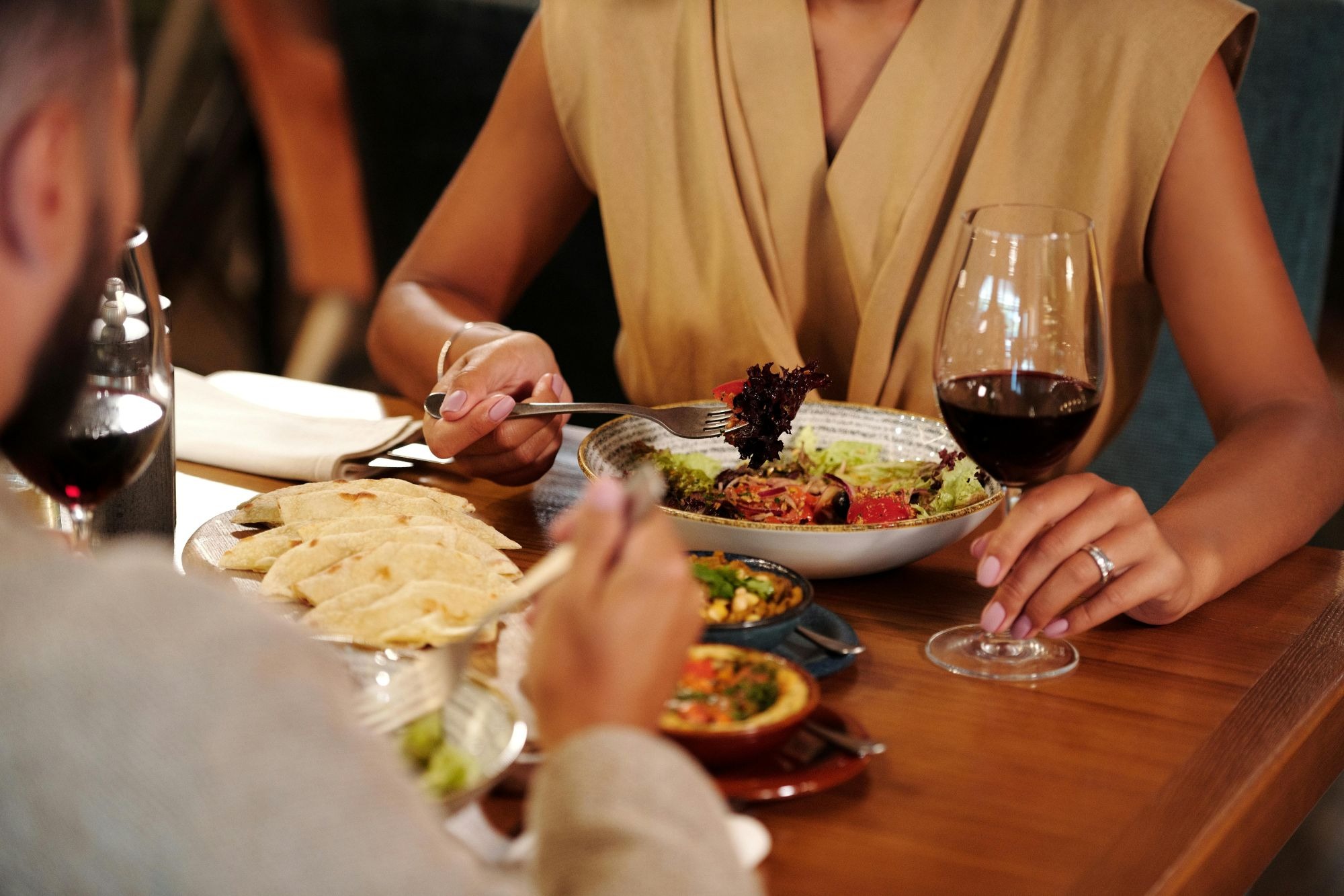 Two people are seated at a wooden table sharing a meal that includes salad, pita bread, and glasses of red wine.