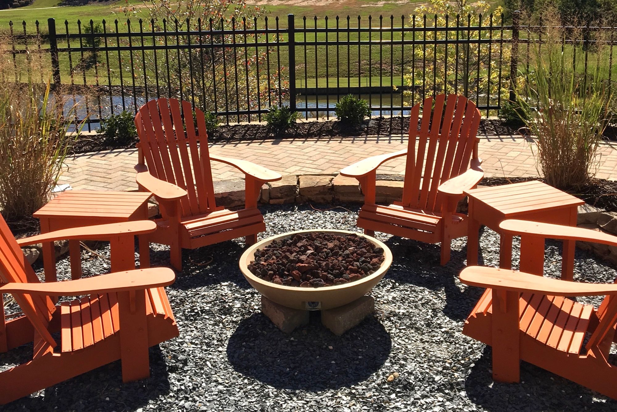 Four orange Adirondack chairs are arranged around a circular stone fire pit filled with dark lava rocks on a gravel surface.
