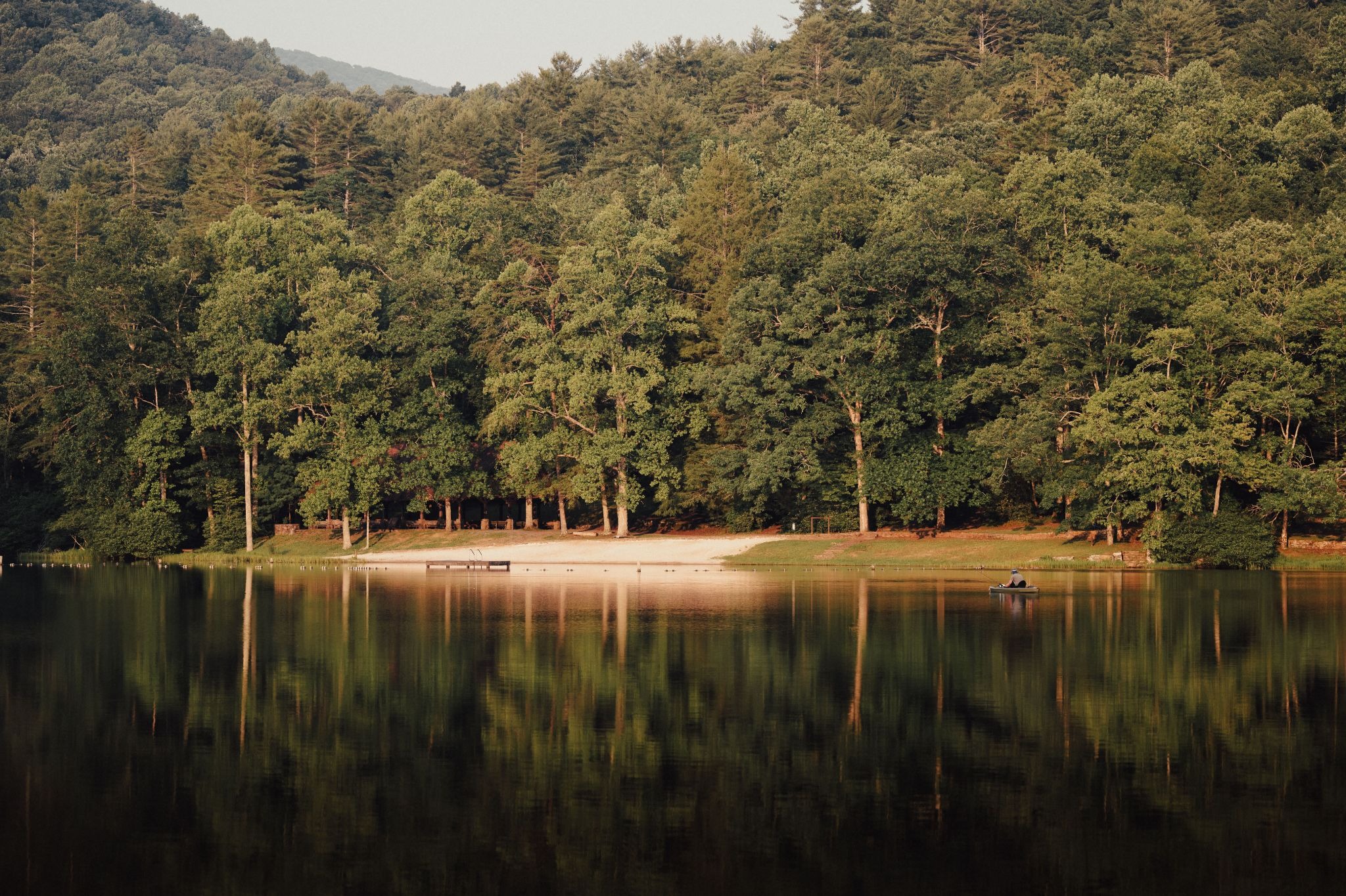 A person paddling a small kayak across a calm mountain lake reflecting a dense green forest and a sandy beach.