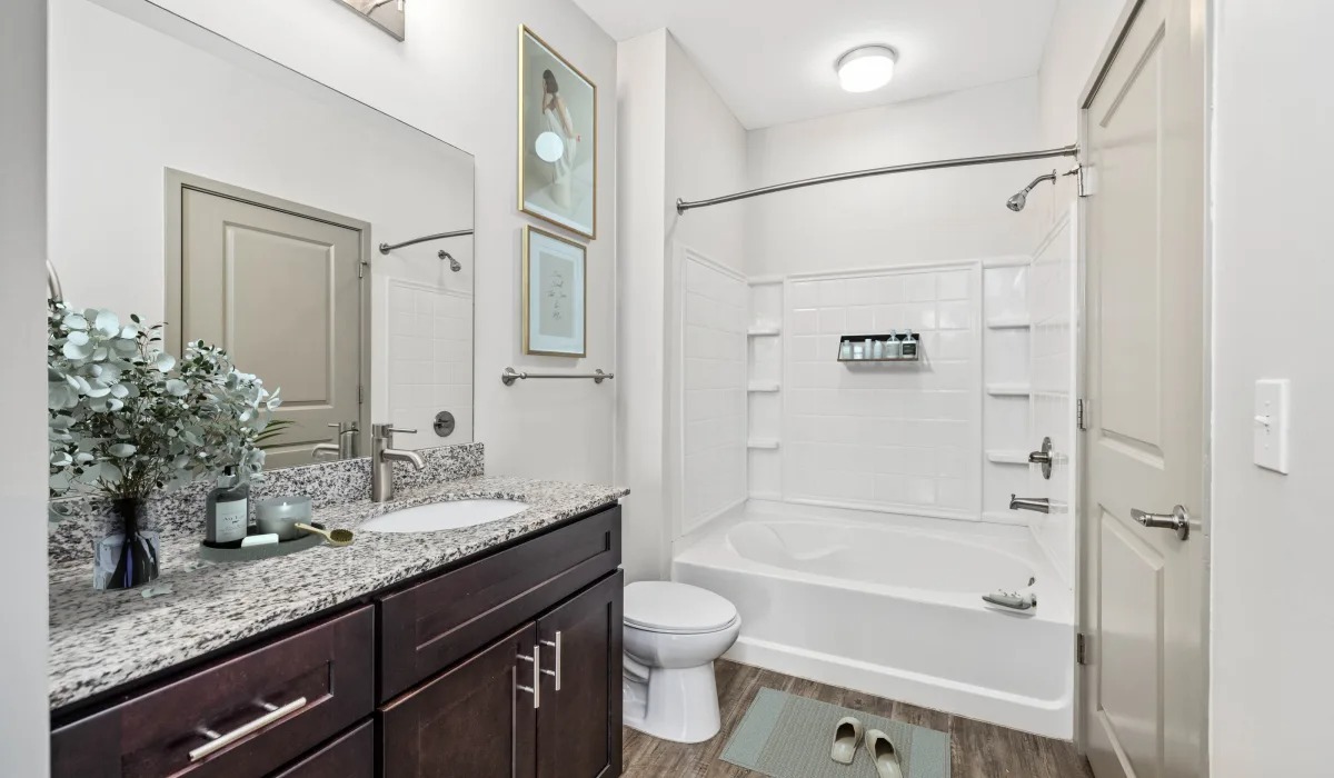 A pristine bathroom showcasing a white shower and tub with dark oak cabinets under a granite countertop that a sink fits onto and a tray with health care items sits.