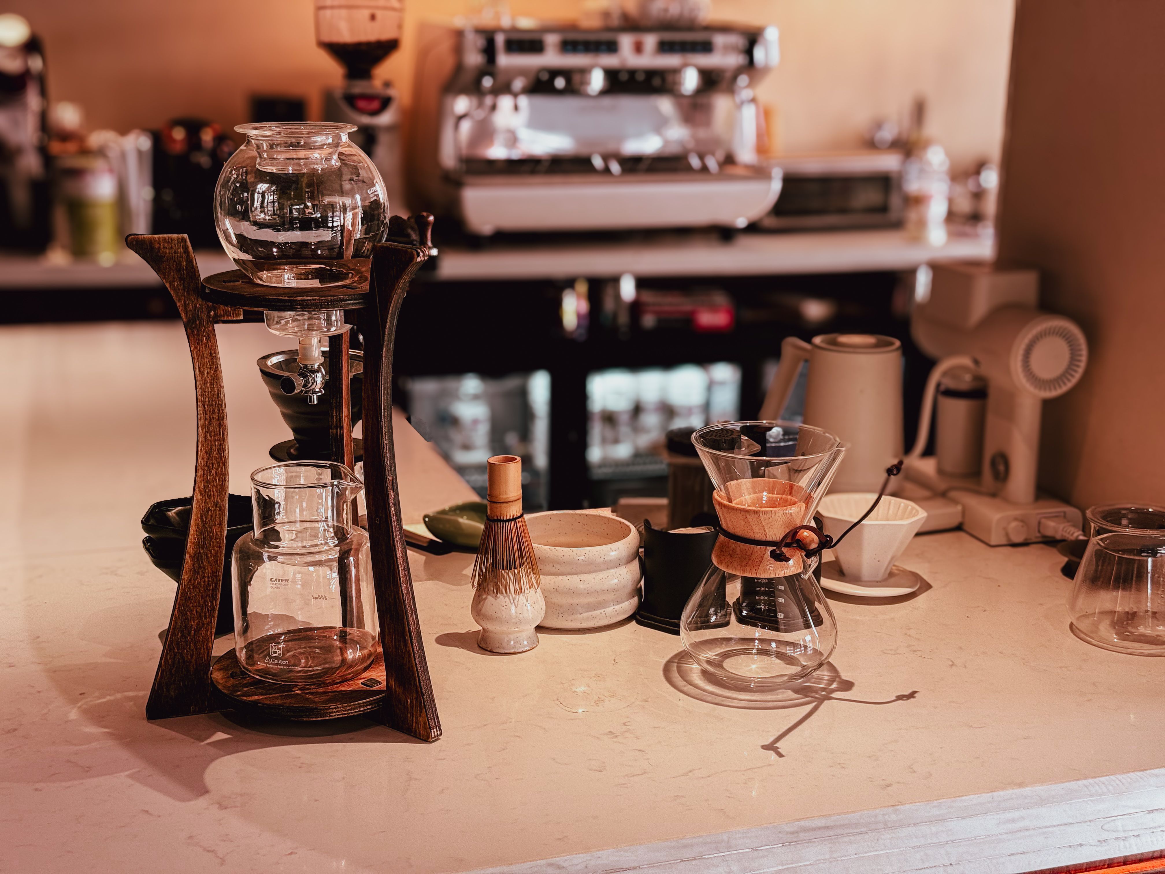 A coffee bar counter displaying specialty equipment including a cold drip tower, Chemex, and a bamboo whisk.