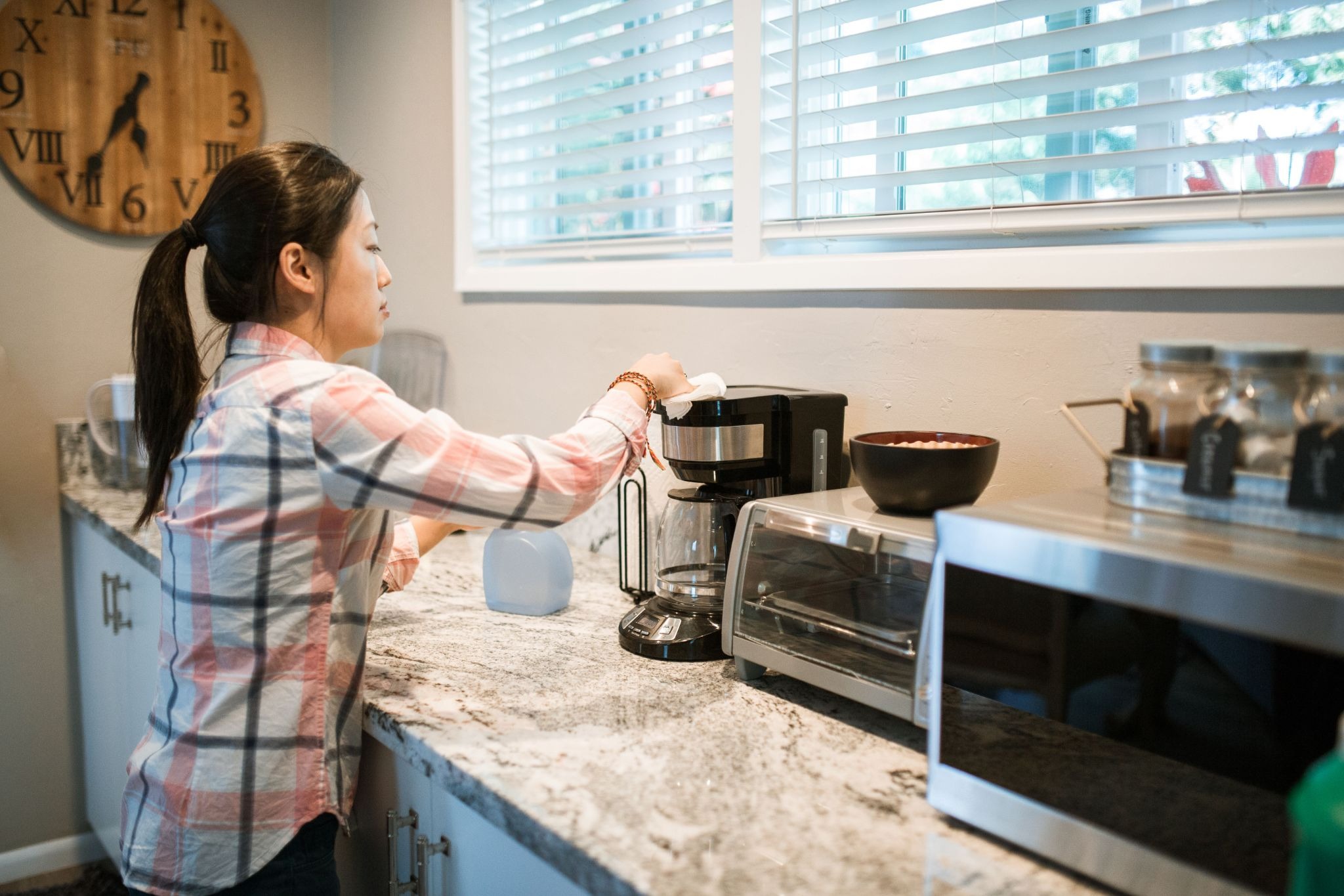 A woman with a ponytail wearing a plaid shirt wiping down a coffee maker on a granite counter in a kitchen.
