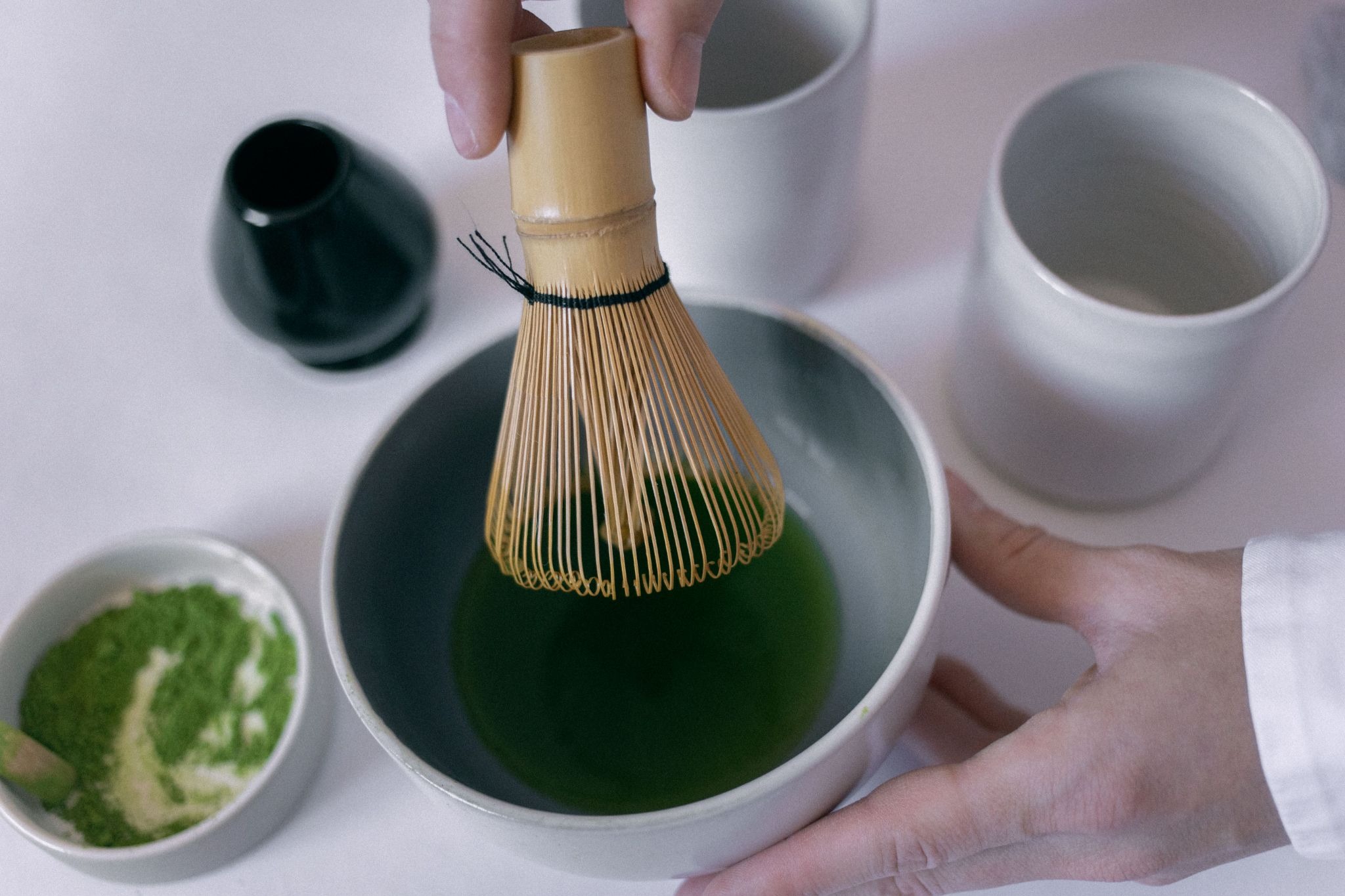 A person using a bamboo whisk to mix green matcha tea inside a grey ceramic bowl on a white table surface.