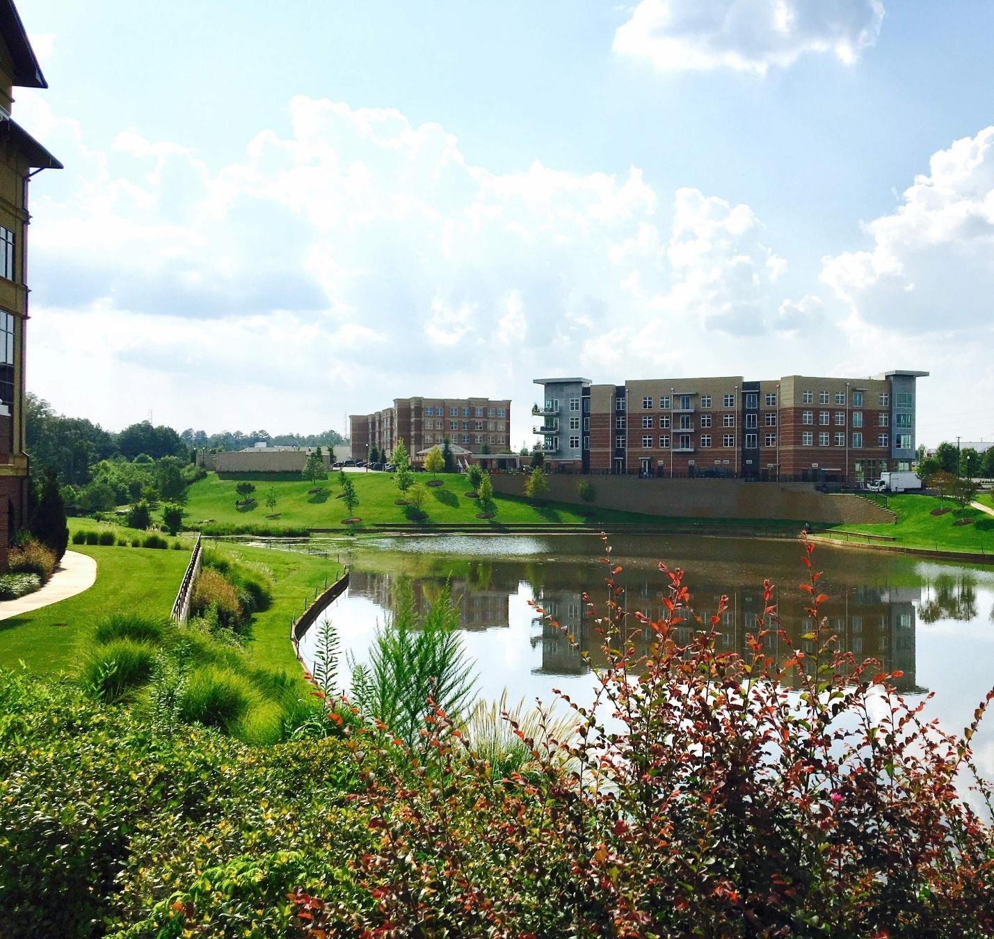 A scenic exterior view of an apartment building reflecting on a calm pond surrounded by lush green landscaping.