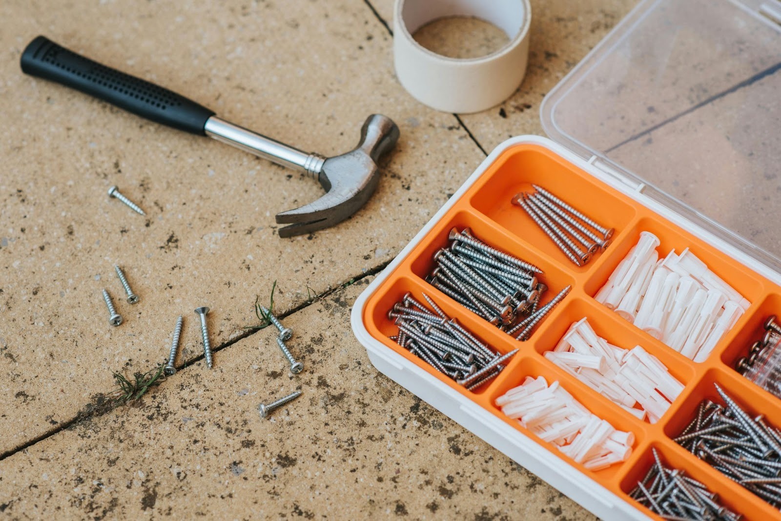 A close-up shows a hammer, loose screws, and an orange parts organizer filled with anchors on a concrete surface.