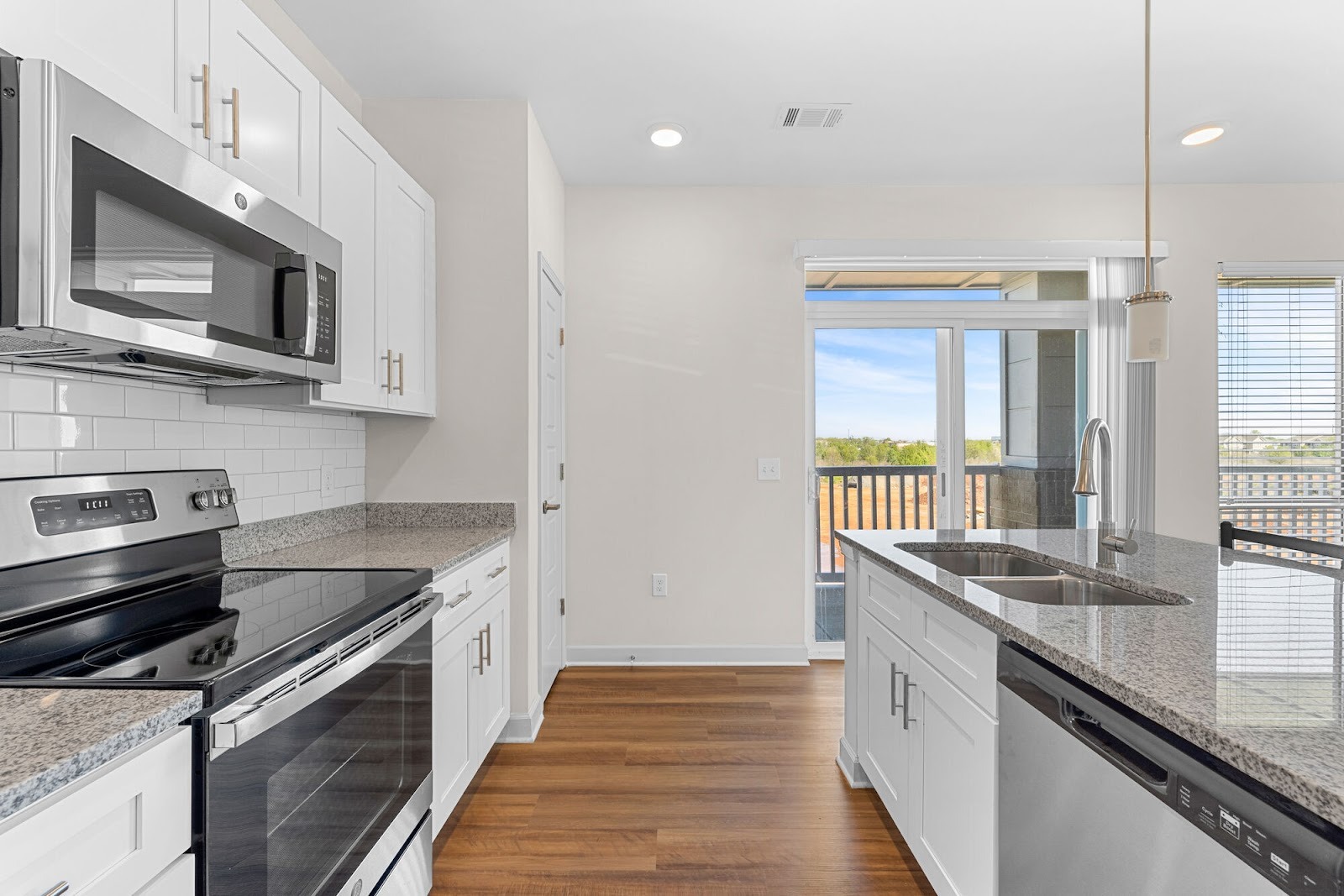 A bright kitchen shows white cabinets, stainless appliances, granite counters, and a sliding door to the balcony.