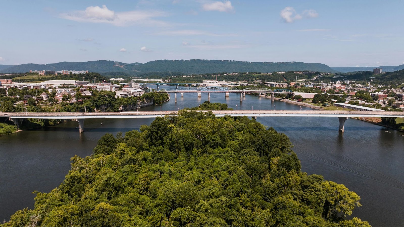 An aerial view of multiple bridges crossing a wide river featuring lush green islands and distant blue mountains.