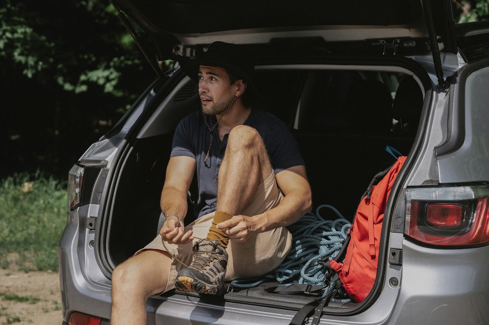 A hiker sitting in the open trunk of a silver SUV tying his boots while preparing for a day of outdoor adventure.