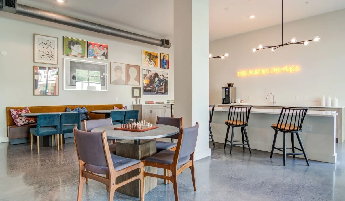 A lounge area with a chess set on a round table, blue chairs, a yellow banquet seat, and a neon sign above a white counter.