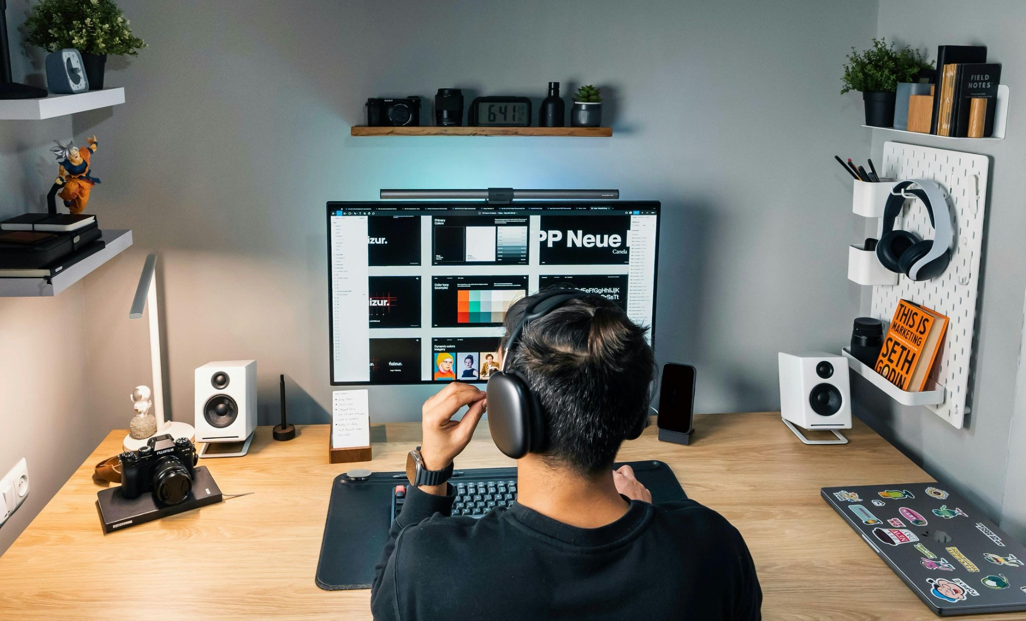 A person viewed from behind wears headphones while sitting at a wooden desk with a large monitor, speakers, and a camera.