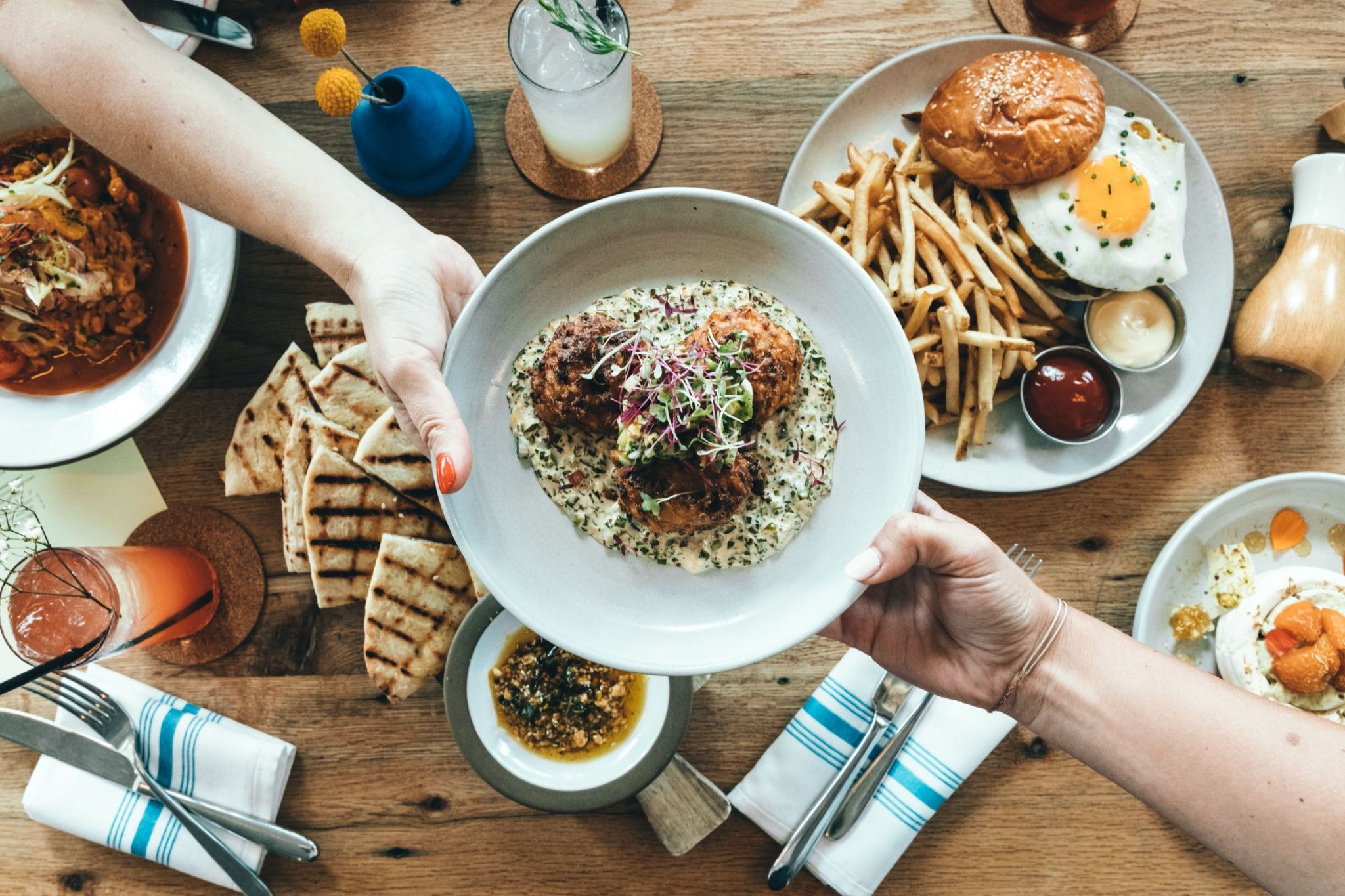 Two people holding a white bowl of food over a wooden dining table featuring a burger, french fries, and cold drinks.