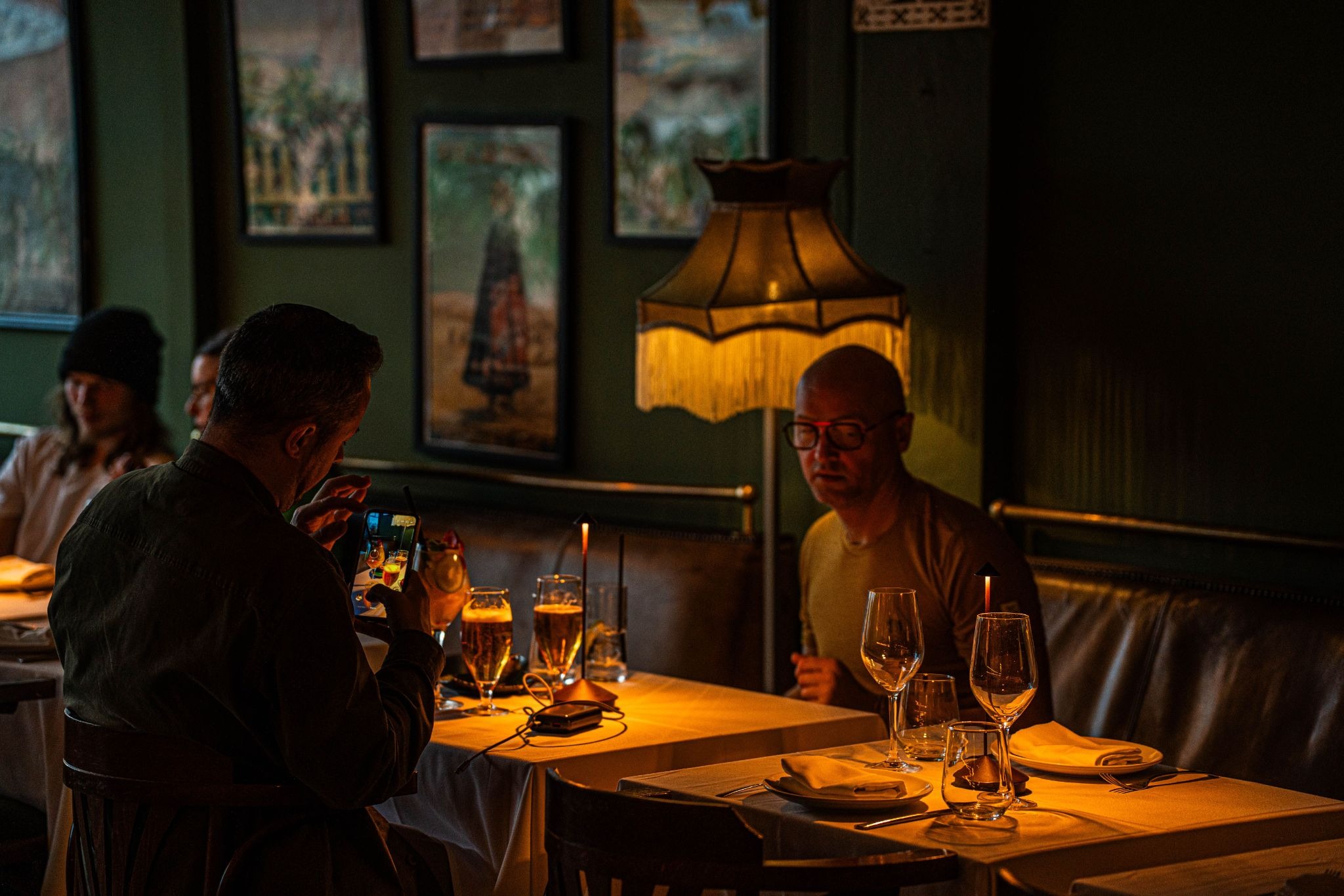 A man sitting at a table inside a dimly lit restaurant using a smartphone and taking a picture of his cocktail glass.