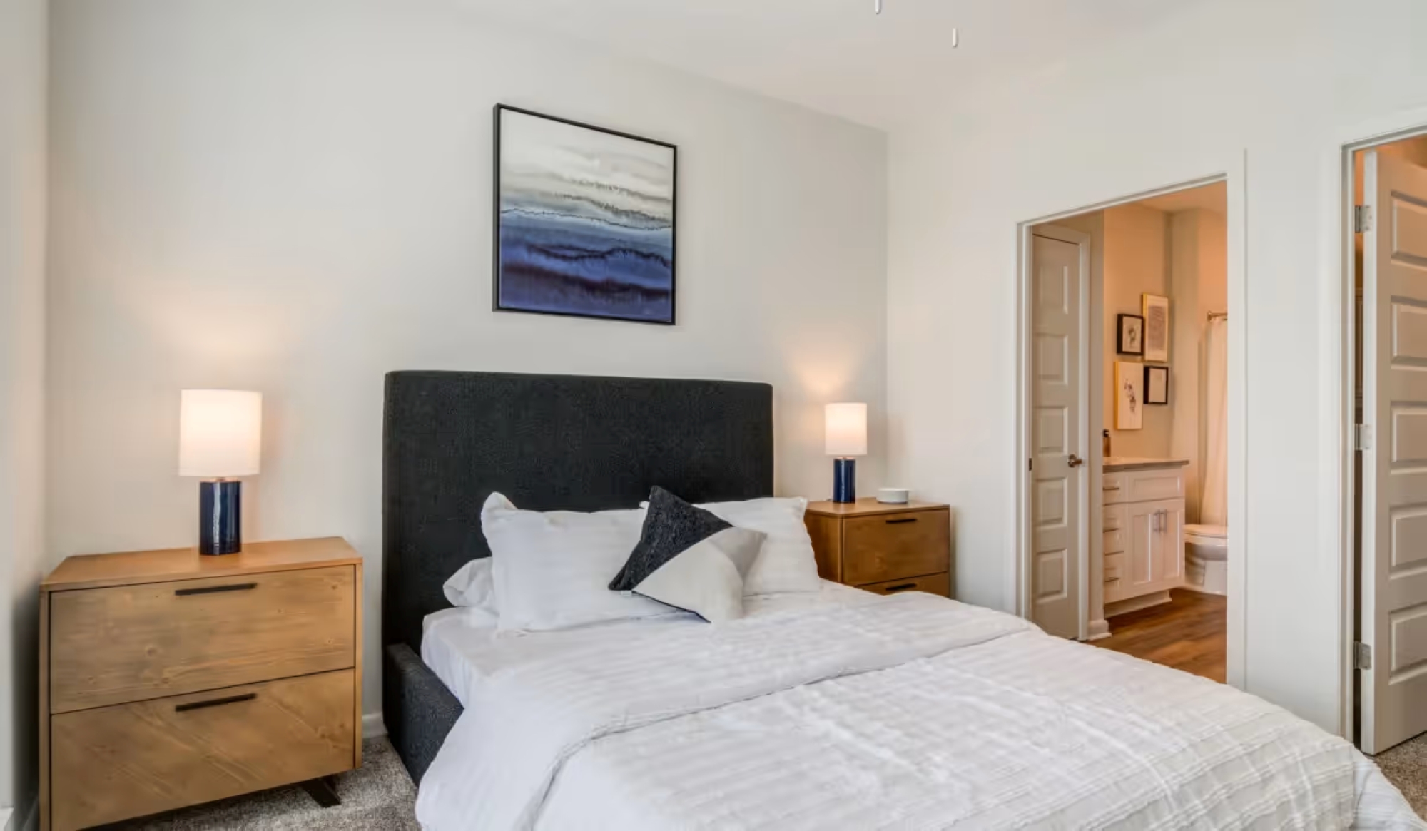 A modern bedroom featuring a dark grey bed frame with white bedding, two wooden nightstands, and a bathroom doorway.