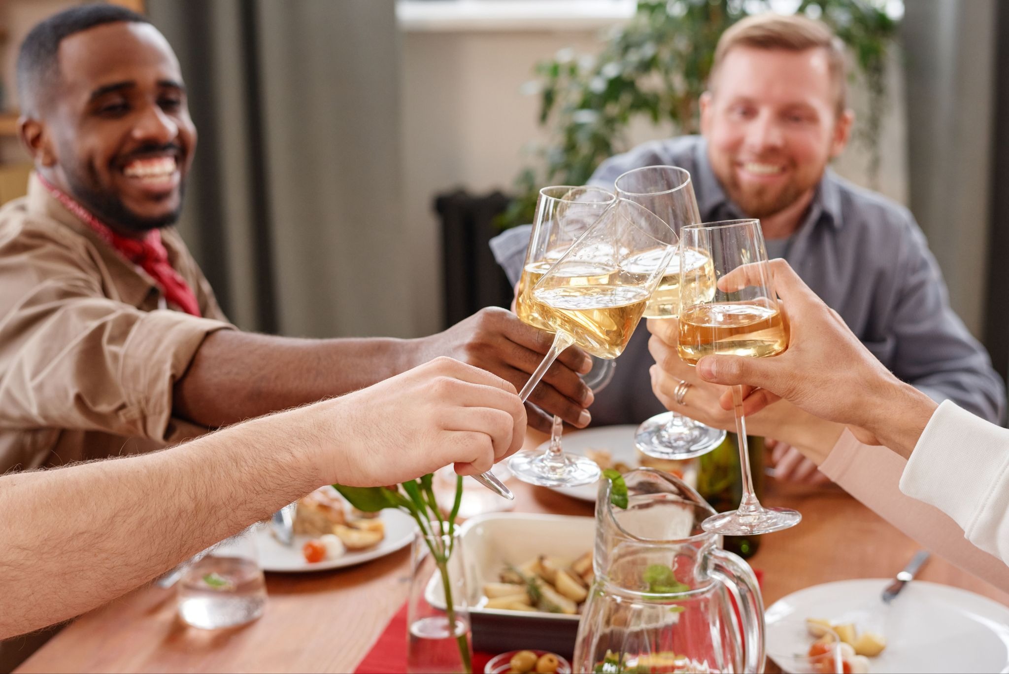 Four people at a dinner party sitting at the dining table cheering their glasses of drinks with food in front of them.