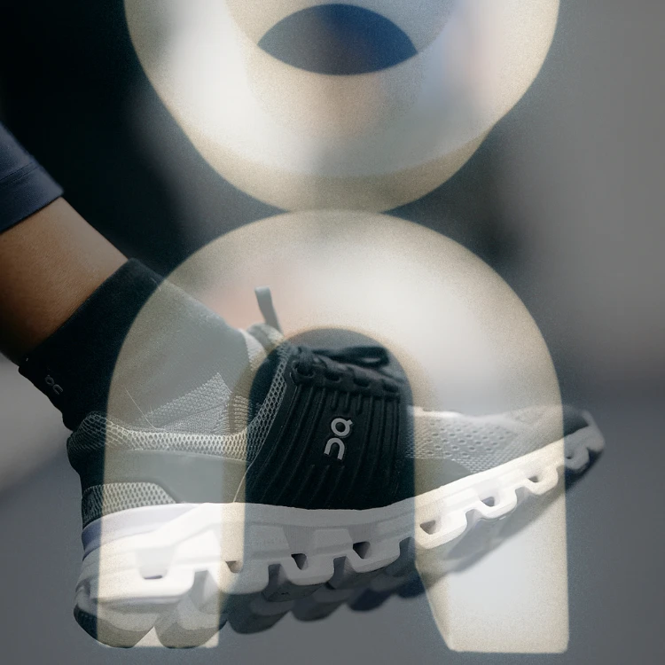 Tênis esportivo preto e cinza com solado branco usando uma meia preta com símbolo branco. (material para empresa ON)