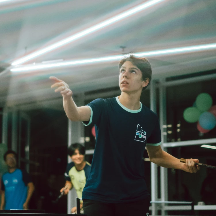 Jovem jogando tênis de mesa em ambiente interno com decoração de balões ao fundo. (materia para empresa Life POng)