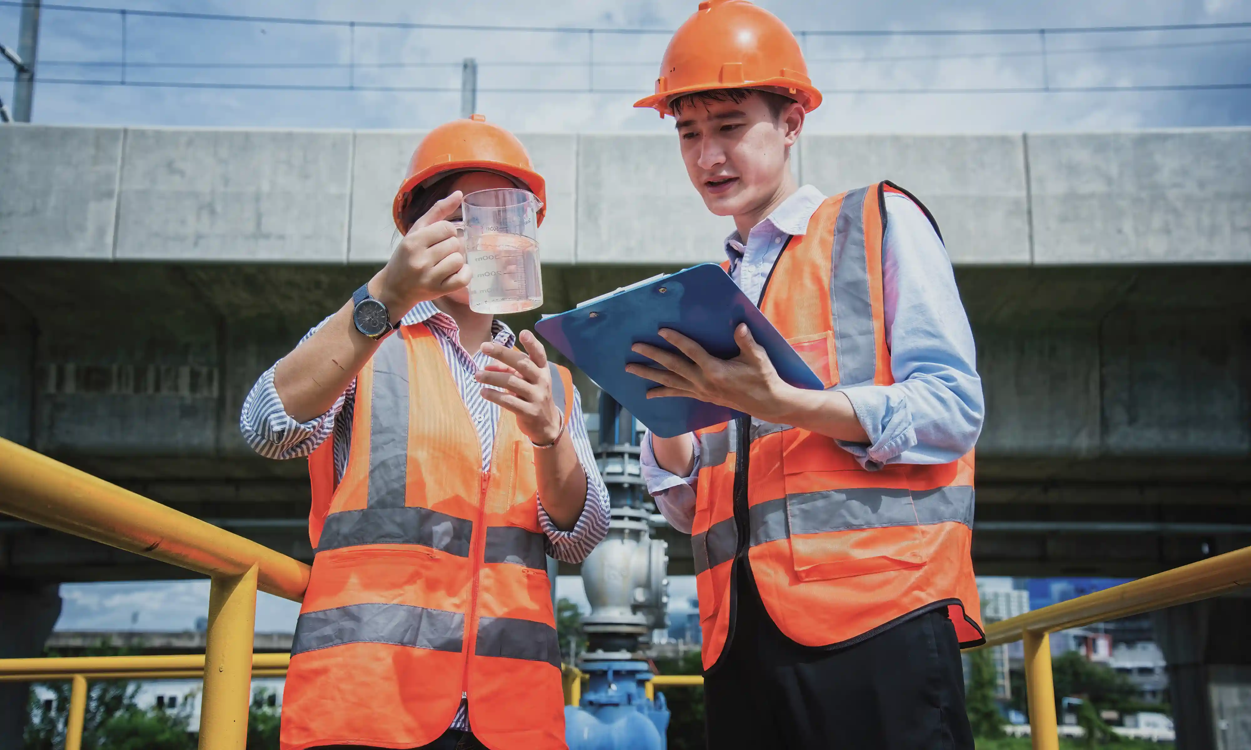 Two construction workers wearing orange safety vests and helmets, one holding a measuring jug with liquid, the other checking notes on a clipboard.