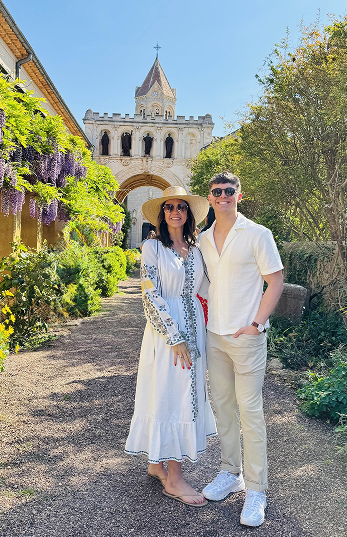 Smiling couple standing on garden path with historic stone building and arch in background under clear blue sky.
