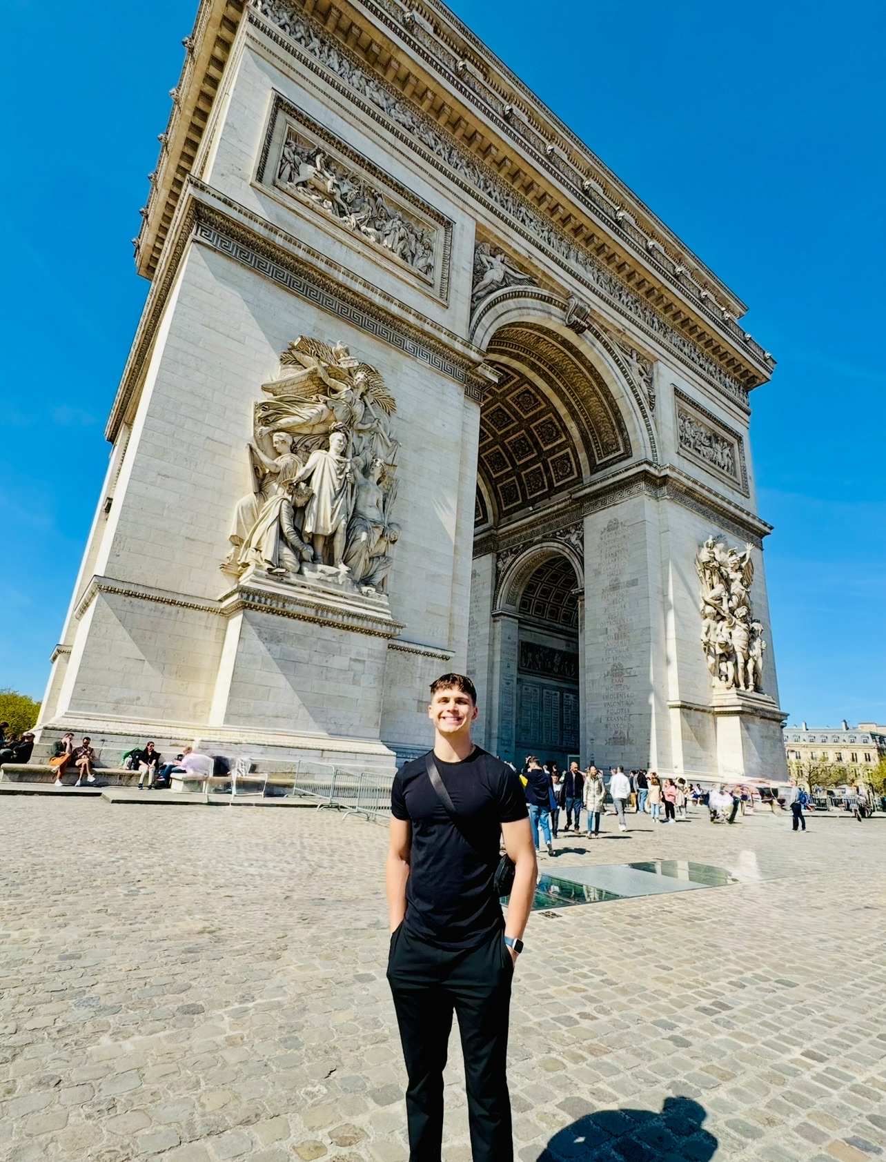Smiling man in black standing in front of the Arc de Triomphe on a sunny day with a clear blue sky.