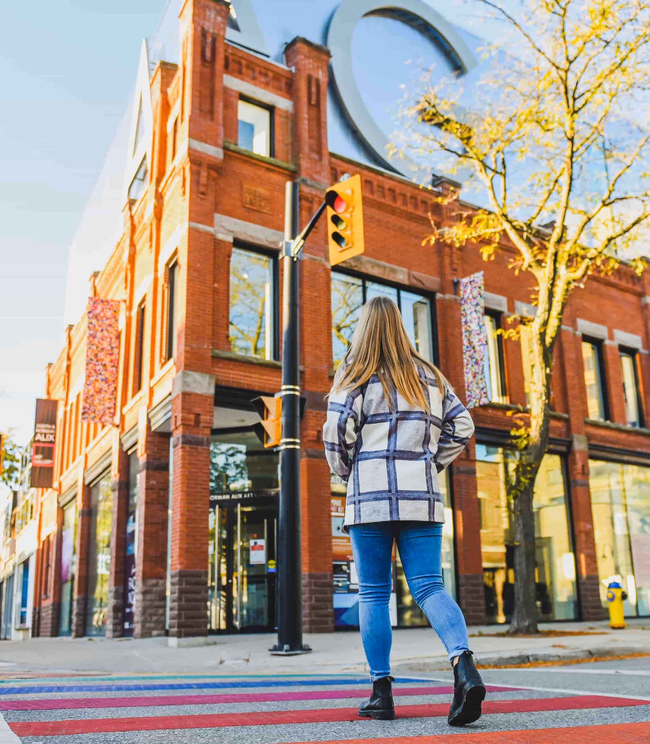 Woman in a plaid jacket and jeans walking across a colorful striped crosswalk near a red brick building and a red traffic light.