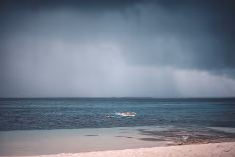 Small wooden boat floating on calm blue sea under dark storm clouds.