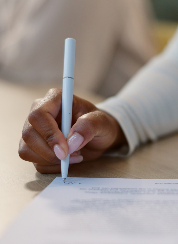 Close-up of a hand holding a white pen and writing on a paper document on a wooden table.