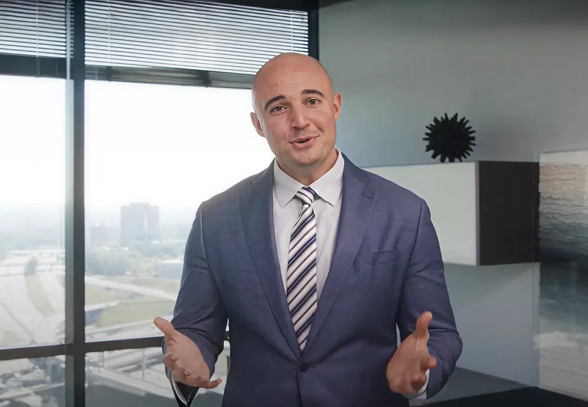 Man in a blue suit and striped tie gesturing with hands in an office with large windows overlooking a city.