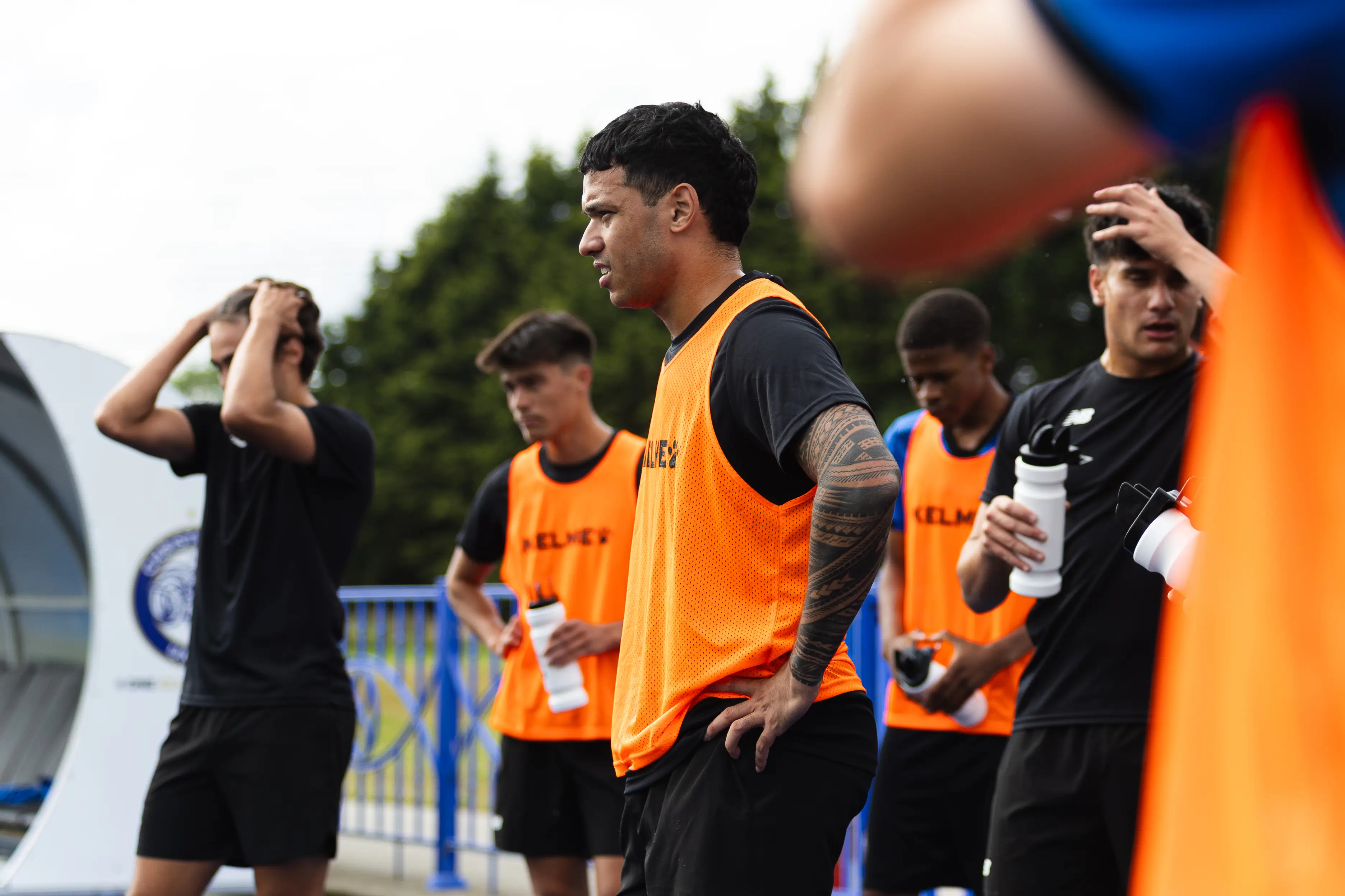Group of young male soccer players in orange training vests taking a break outdoors, some holding water bottles.