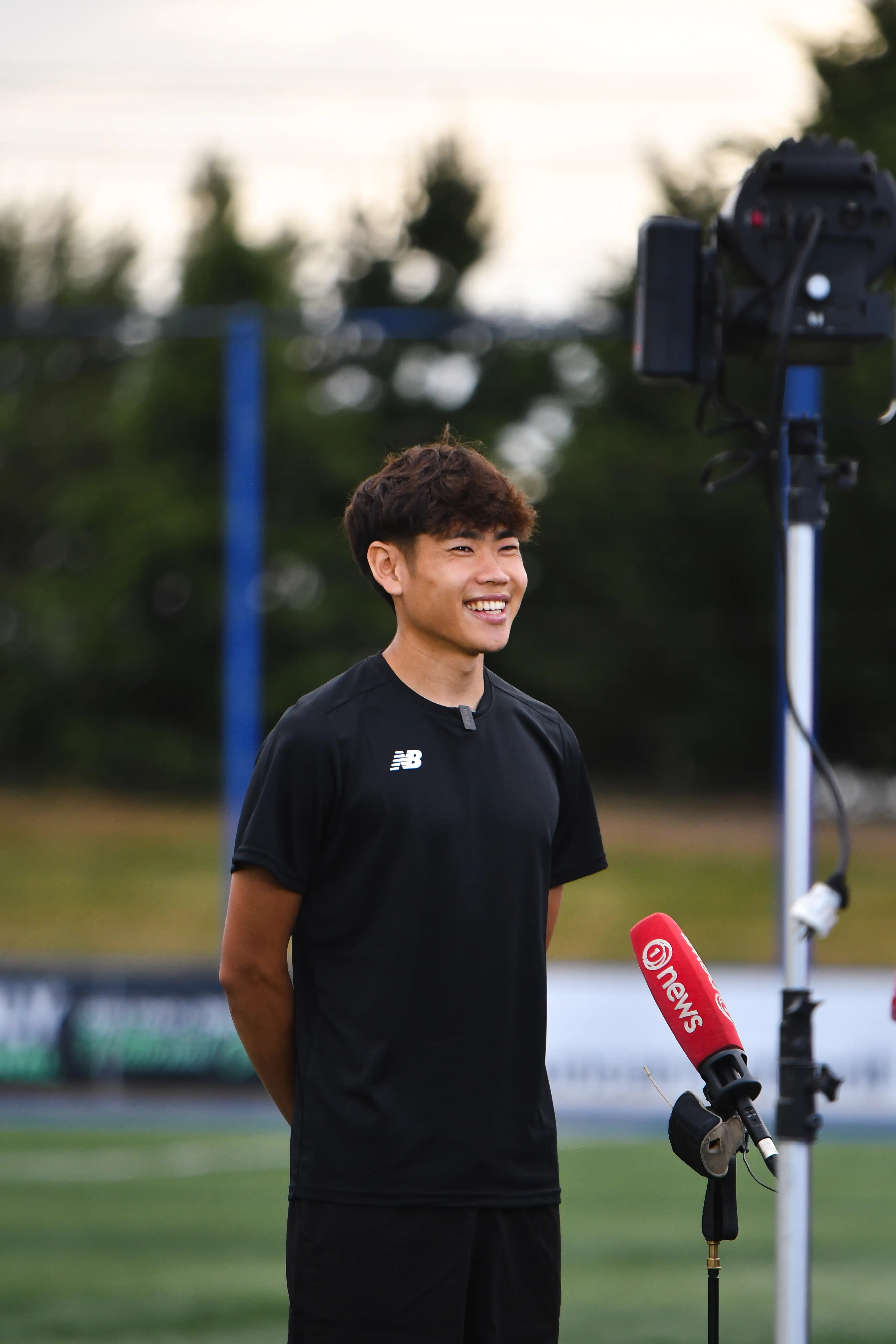 Young man in black sportswear smiling during an outdoor interview with a red microphone labeled 'news'.