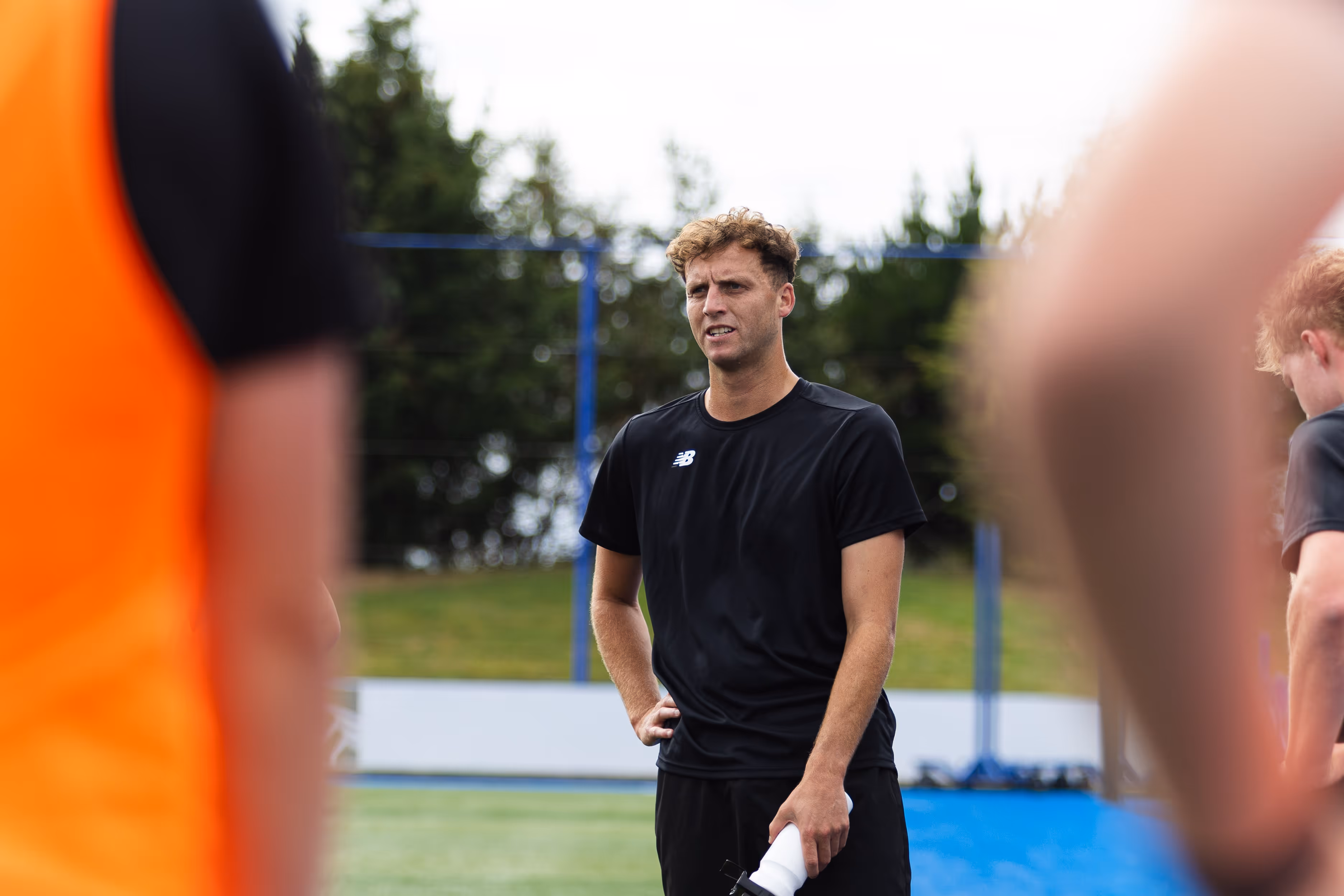 Man in black sportswear holding a water bottle standing on a sports field during practice.