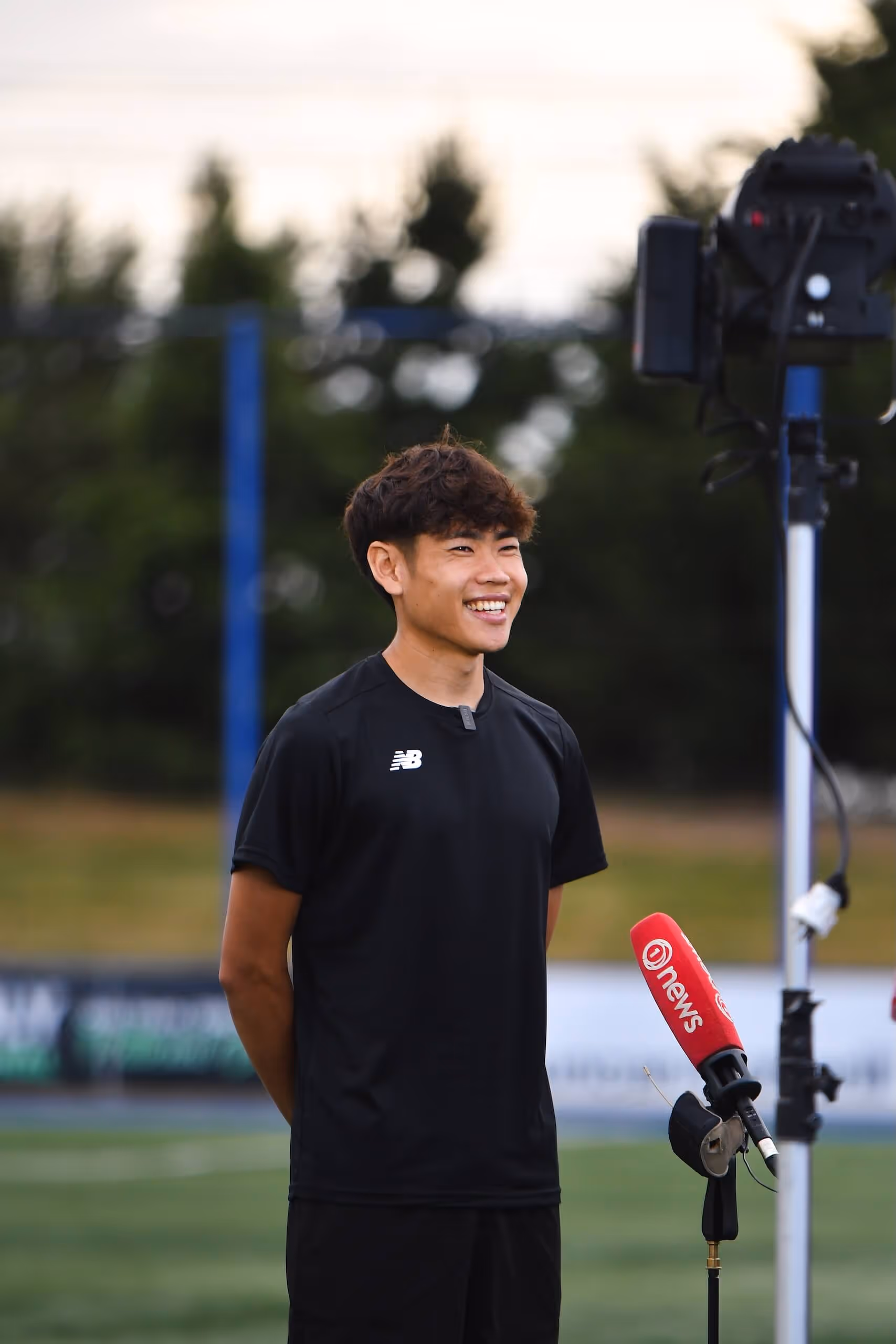 Young man in black sportswear smiling during an outdoor interview with a red microphone labeled 'news'.