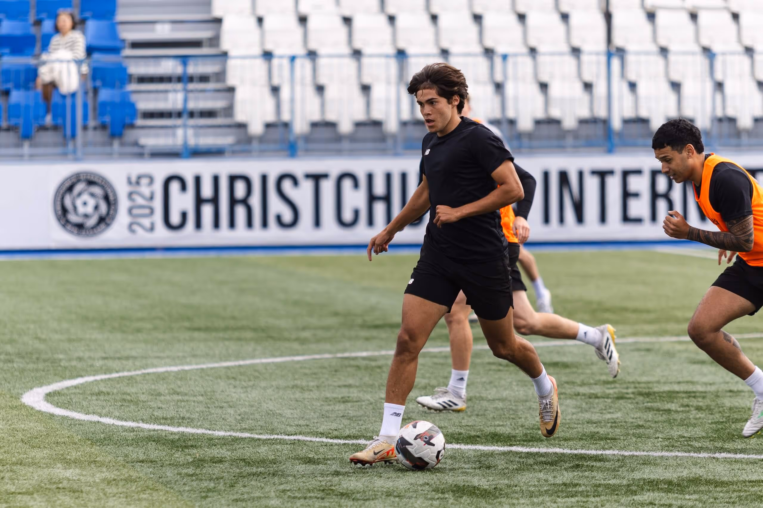 Soccer player in black controlling the ball on a field while two players in orange vests chase him.