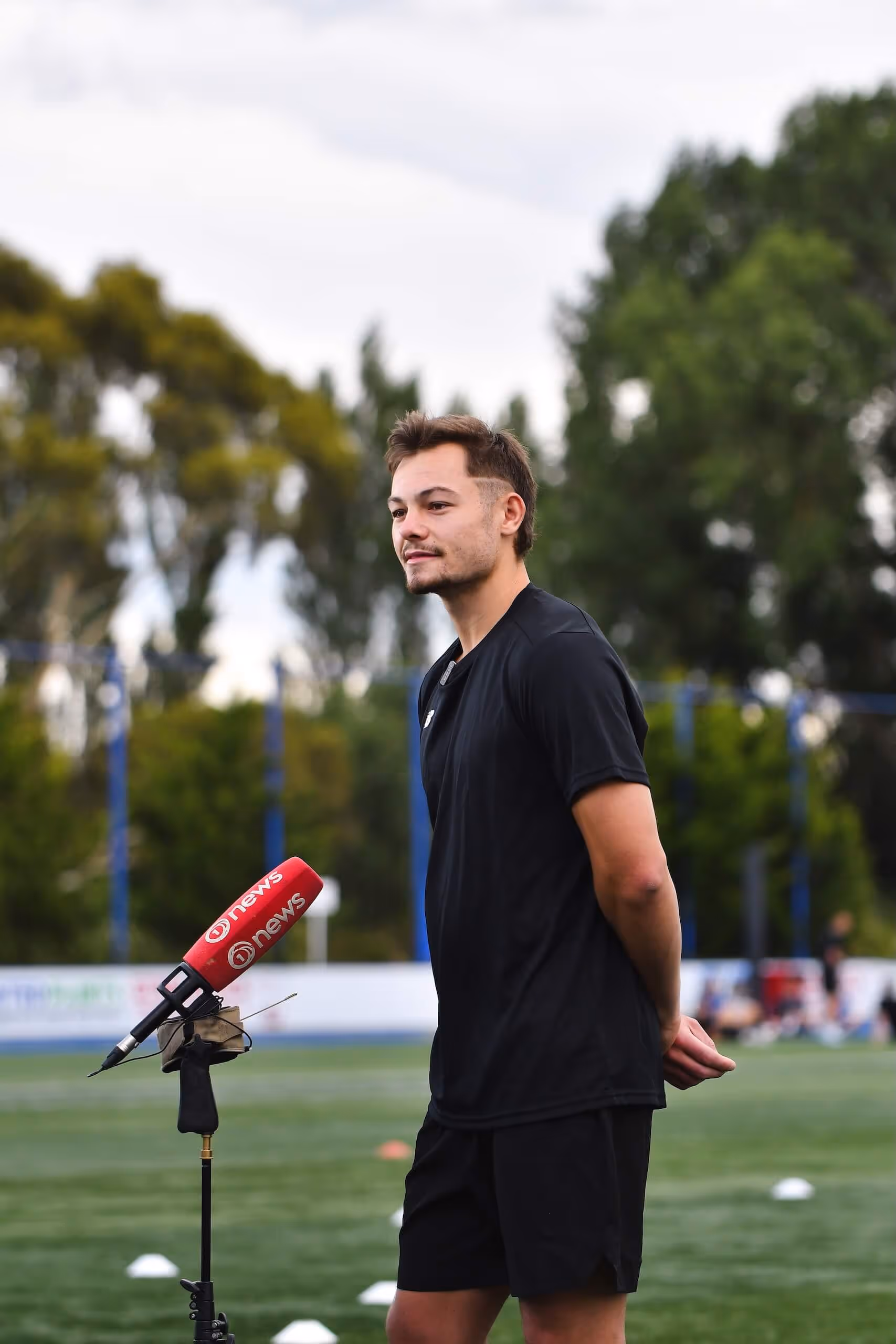 Young man in black sportswear standing with hands behind back facing a microphone on a soccer field.