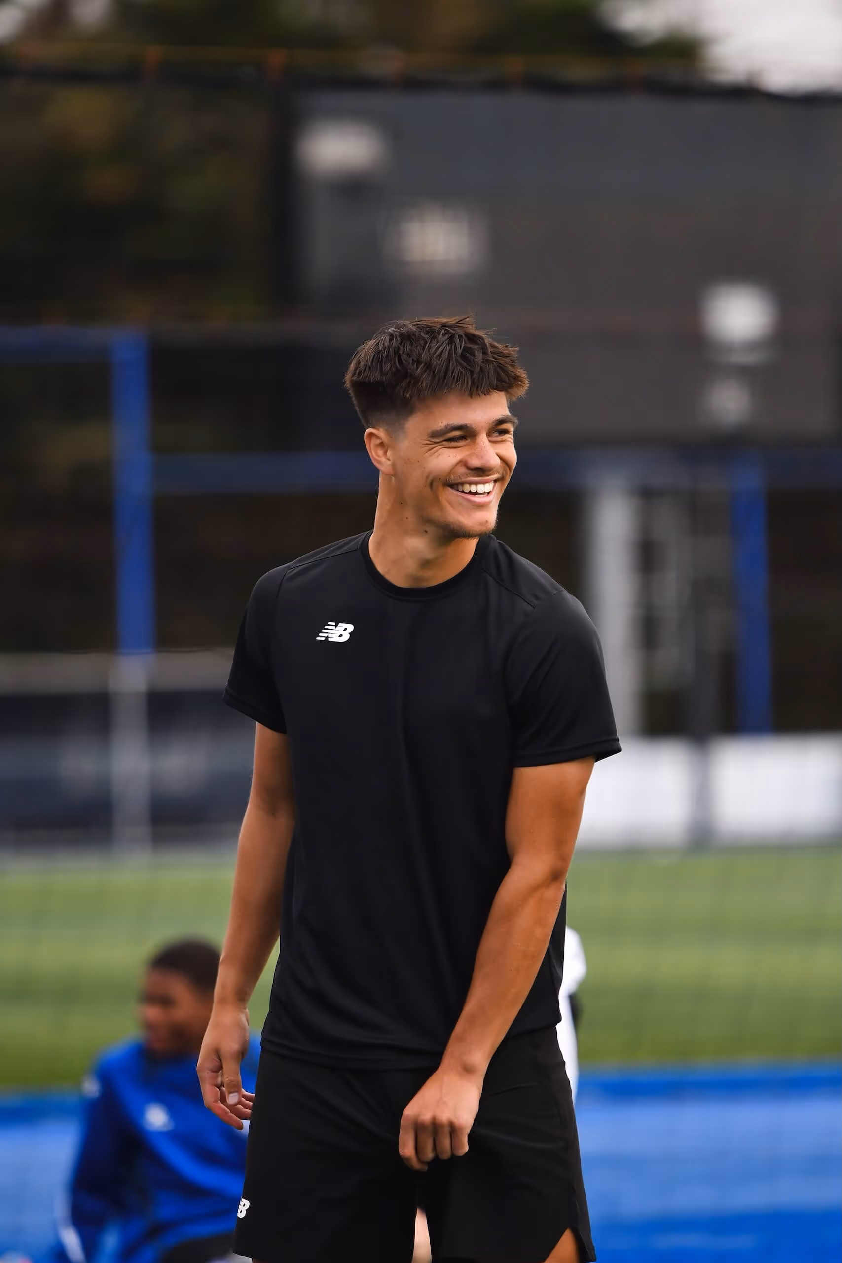 Smiling young man in black athletic wear standing on a sports field with a blurred player in blue in the background.