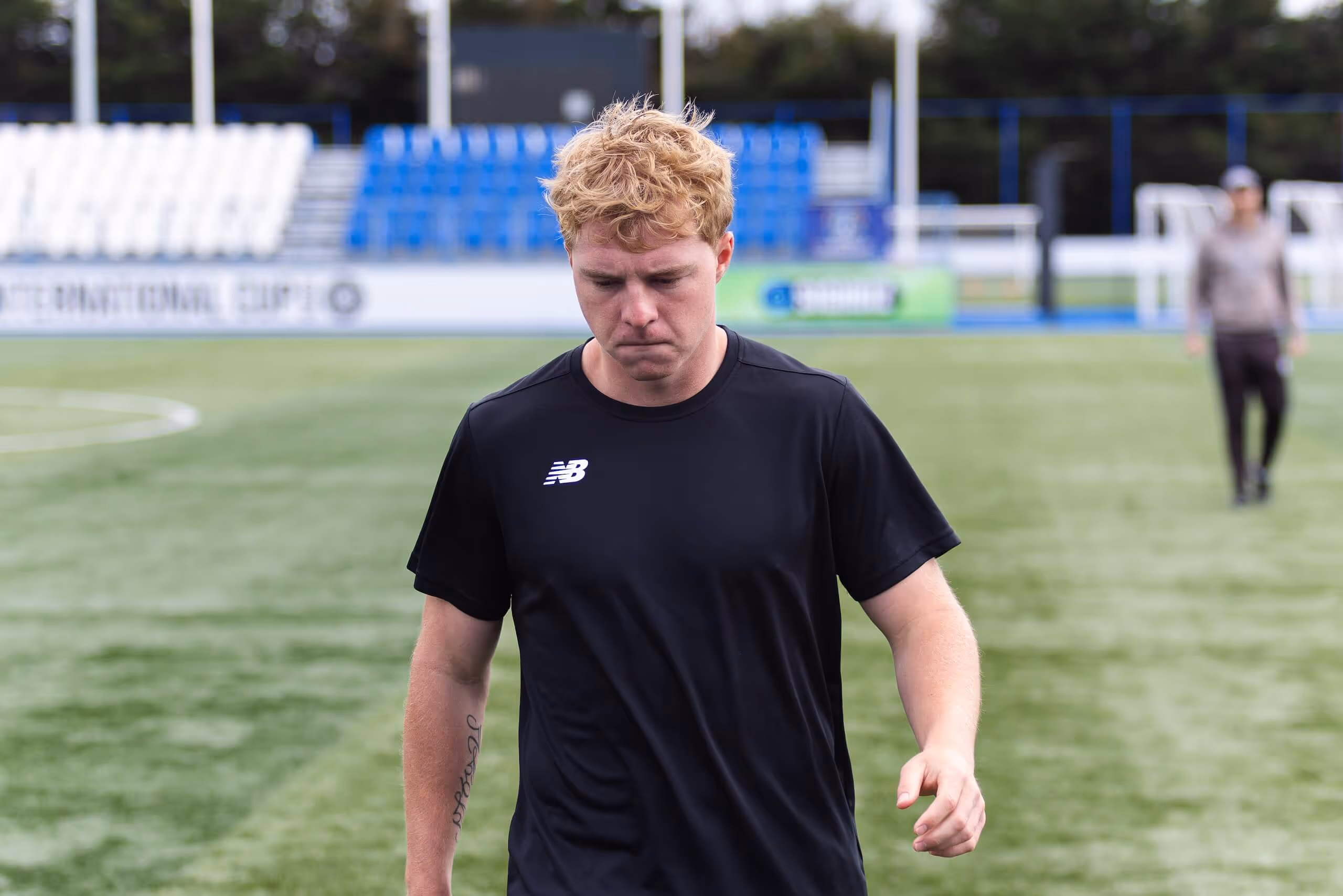 Young man with curly blond hair wearing a black New Balance shirt walking on a soccer field with empty blue and white stadium seats in the background.