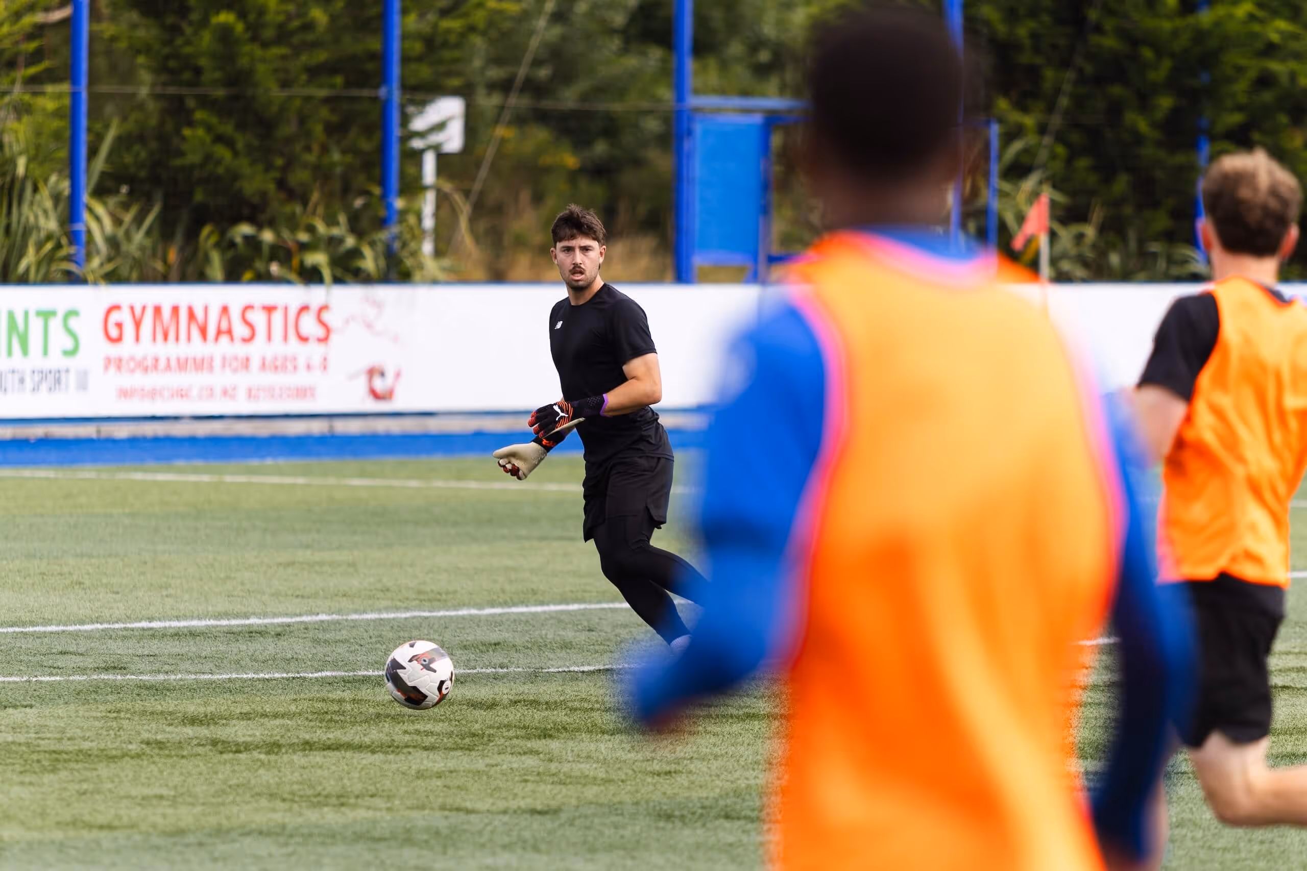 Soccer goalkeeper in black uniform preparing to kick a ball on the field while two players in orange training vests look on.