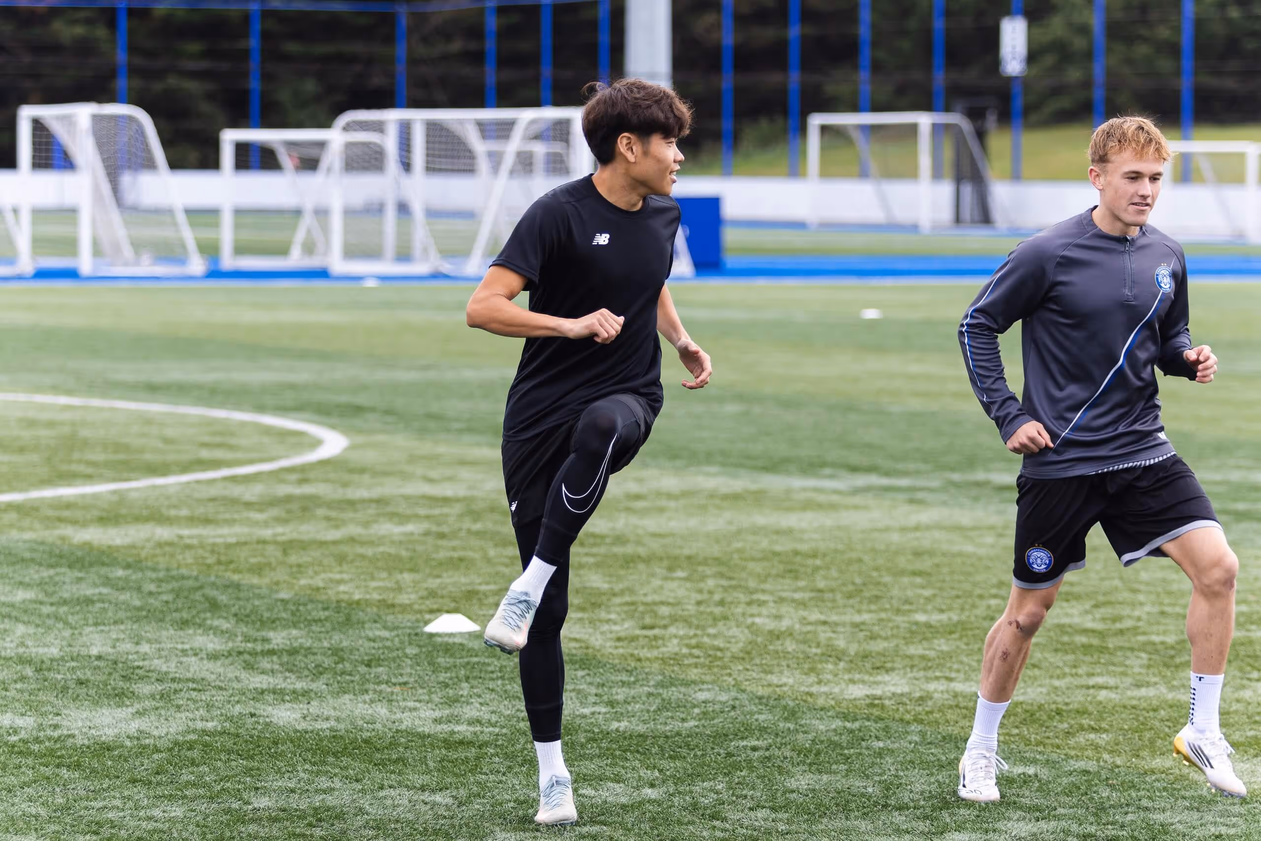 Two men warming up and jogging on a soccer field with multiple goal nets in the background.