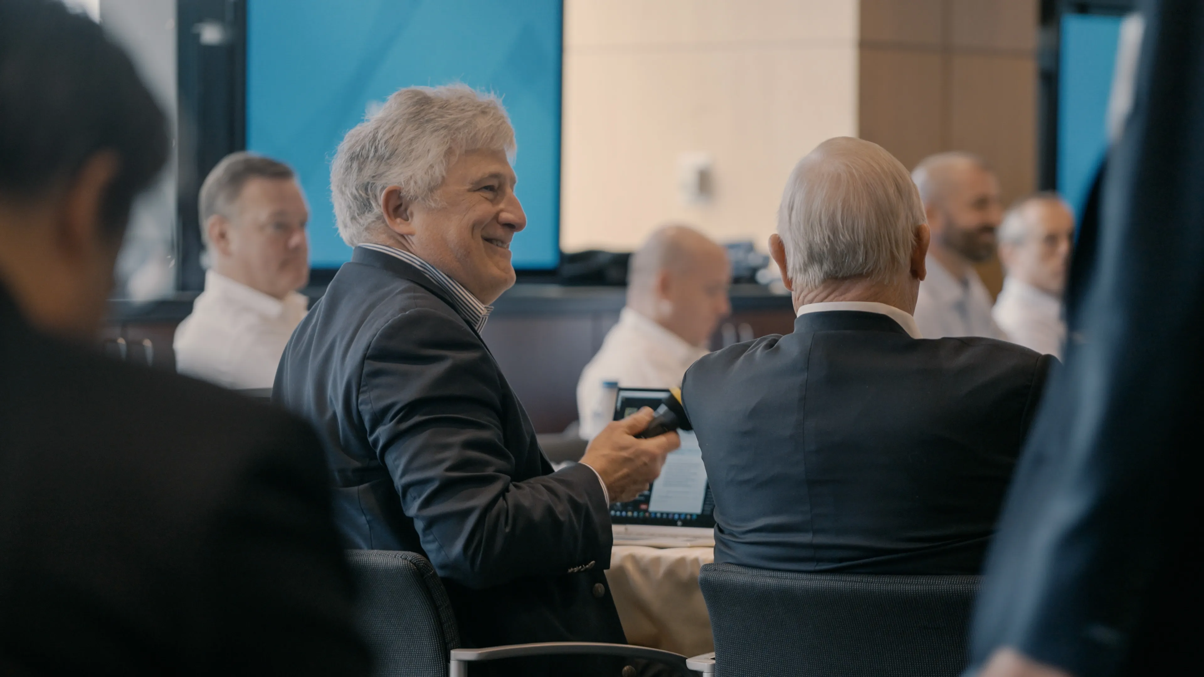 A group of men in business attire seated around a conference table during a meeting, one smiling and holding a remote.